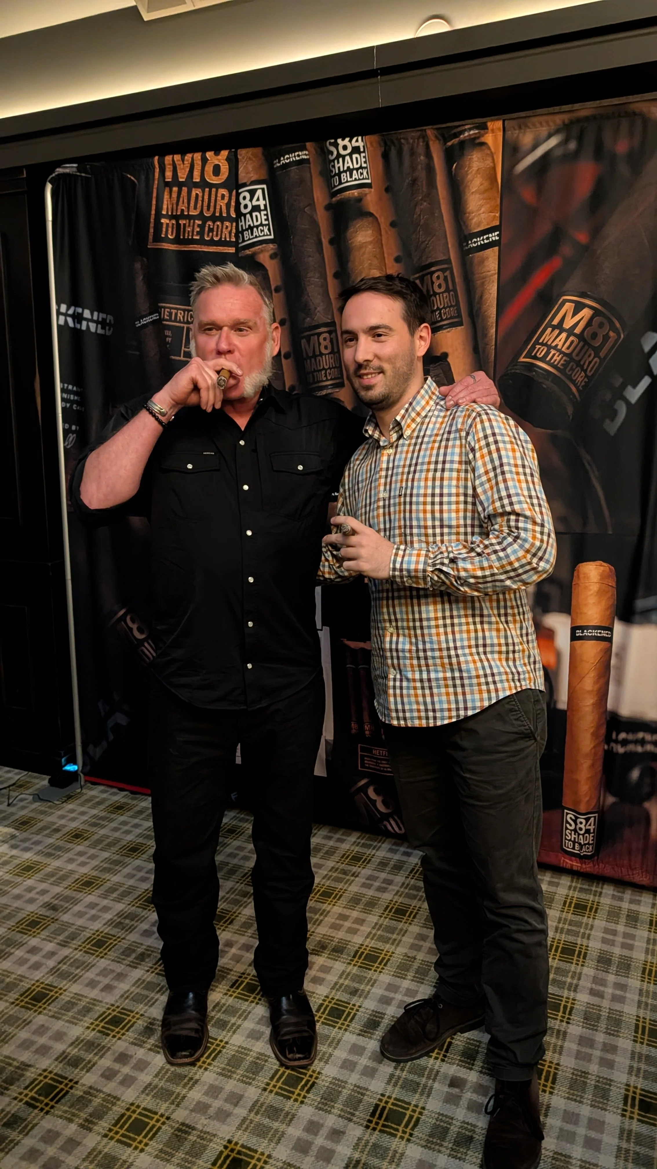 Two men posing together in front of a black backdrop with large cigar images and text. One man has gray hair, a beard, and is smoking a cigar. Both are smiling and holding cigars.