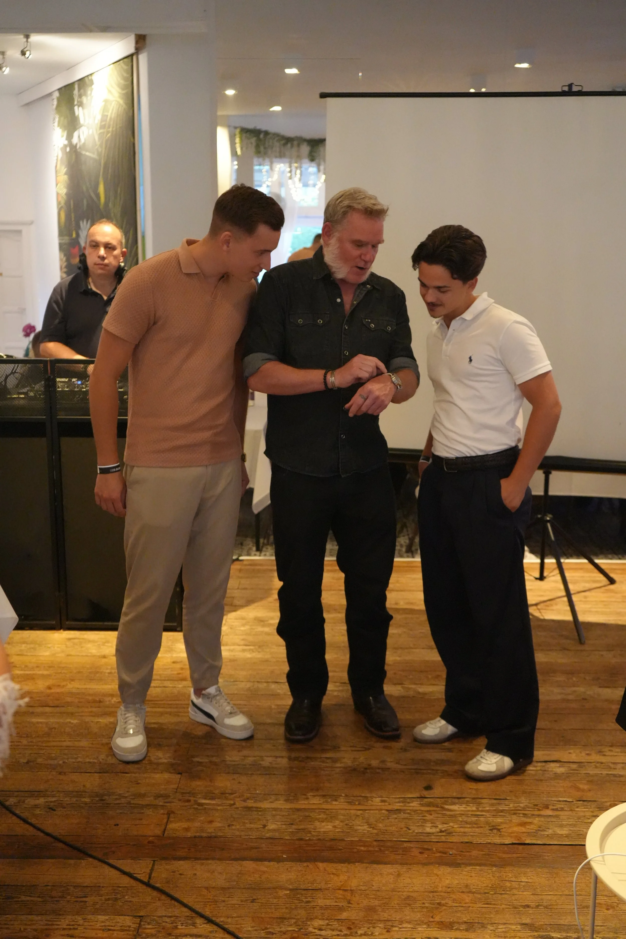 Four men at a social gathering, three are standing close together and looking at a wristwatch, one is in the background near a DJ setup.