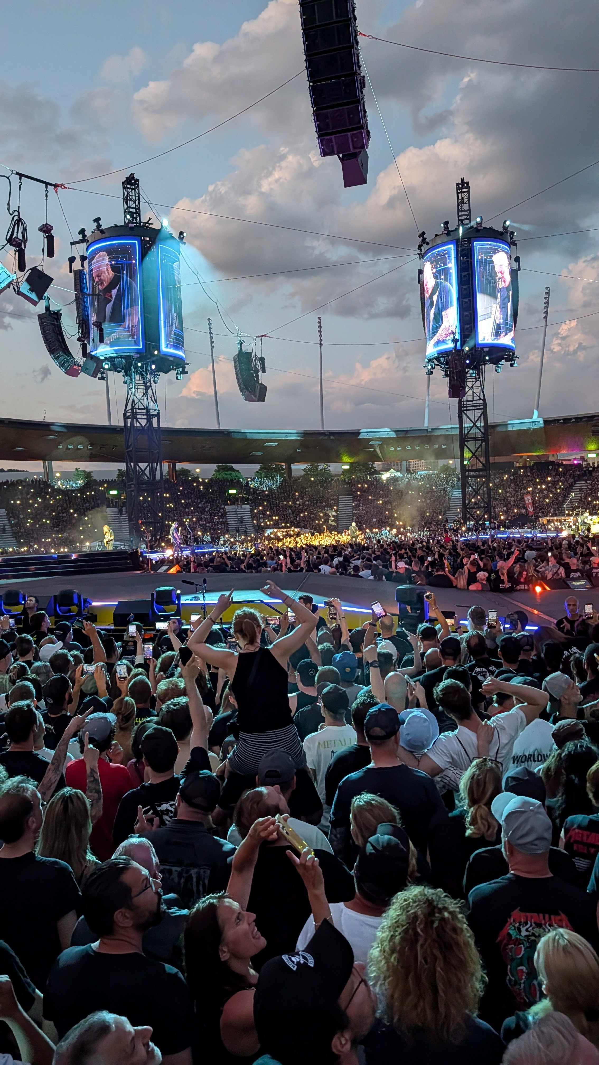 Crowd of people attending a Metallica concert at an outdoor venue with two large screens showing a performer, stage lighting, and a cloudy sky.