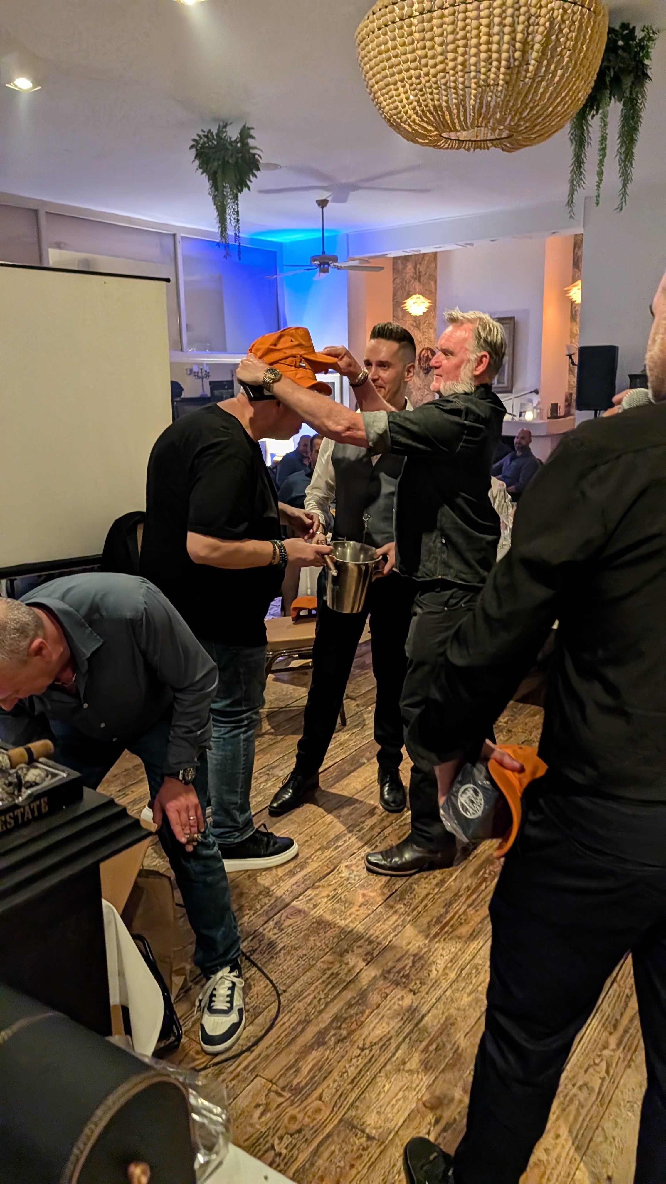 A group of people at a social gathering, with one man placing an orange hat on another person's head. The setting appears to be indoors, possibly a restaurant or bar, with warm lighting, wooden flooring, and hanging plants.