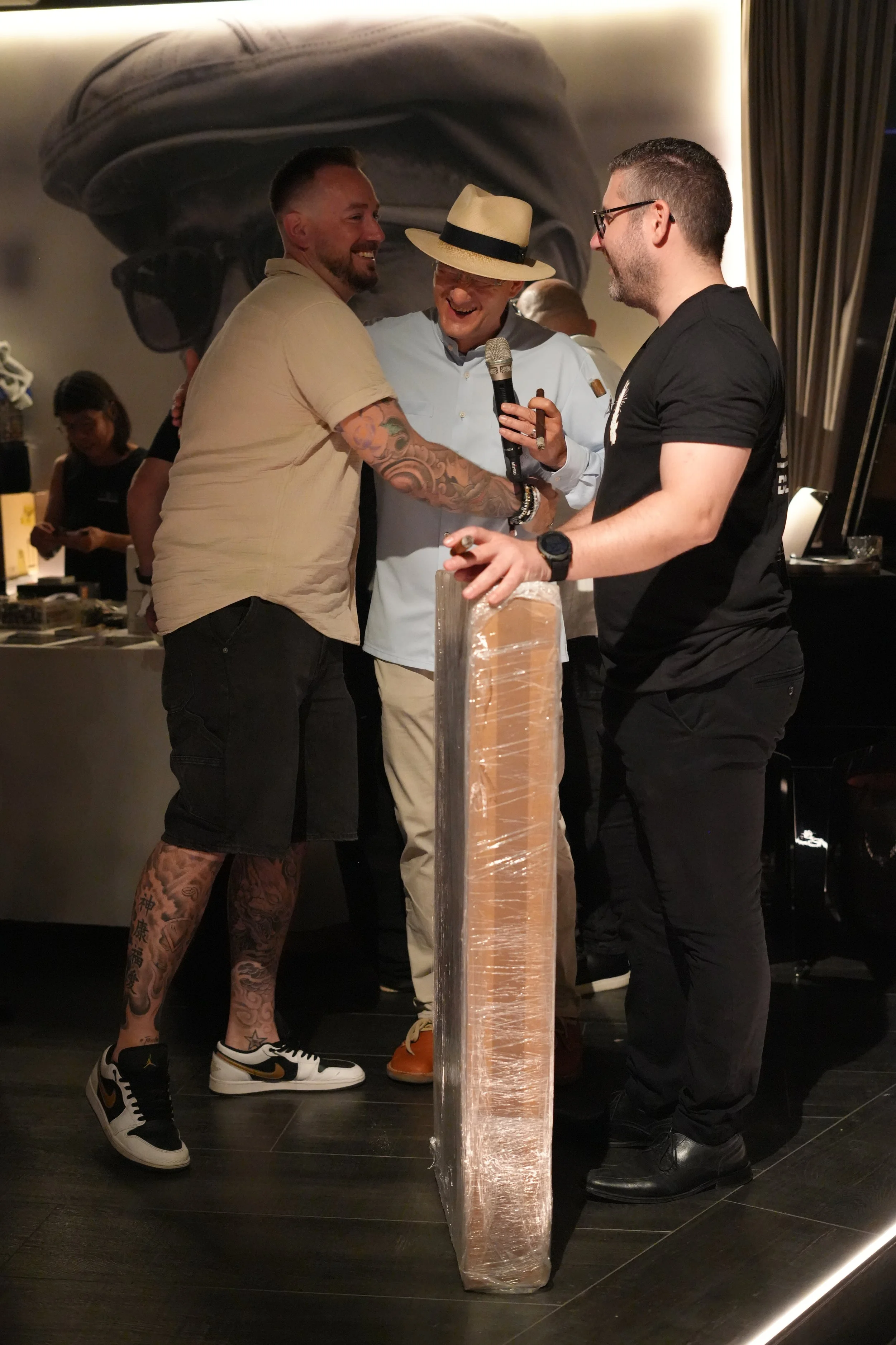 Three men are smiling and talking in a room with a mural of sunglasses on the wall behind them. One man is wearing a beige shirt, black shorts, and sneakers, has tattoos on his legs, and is holding hands with another man. The second man wears a light
