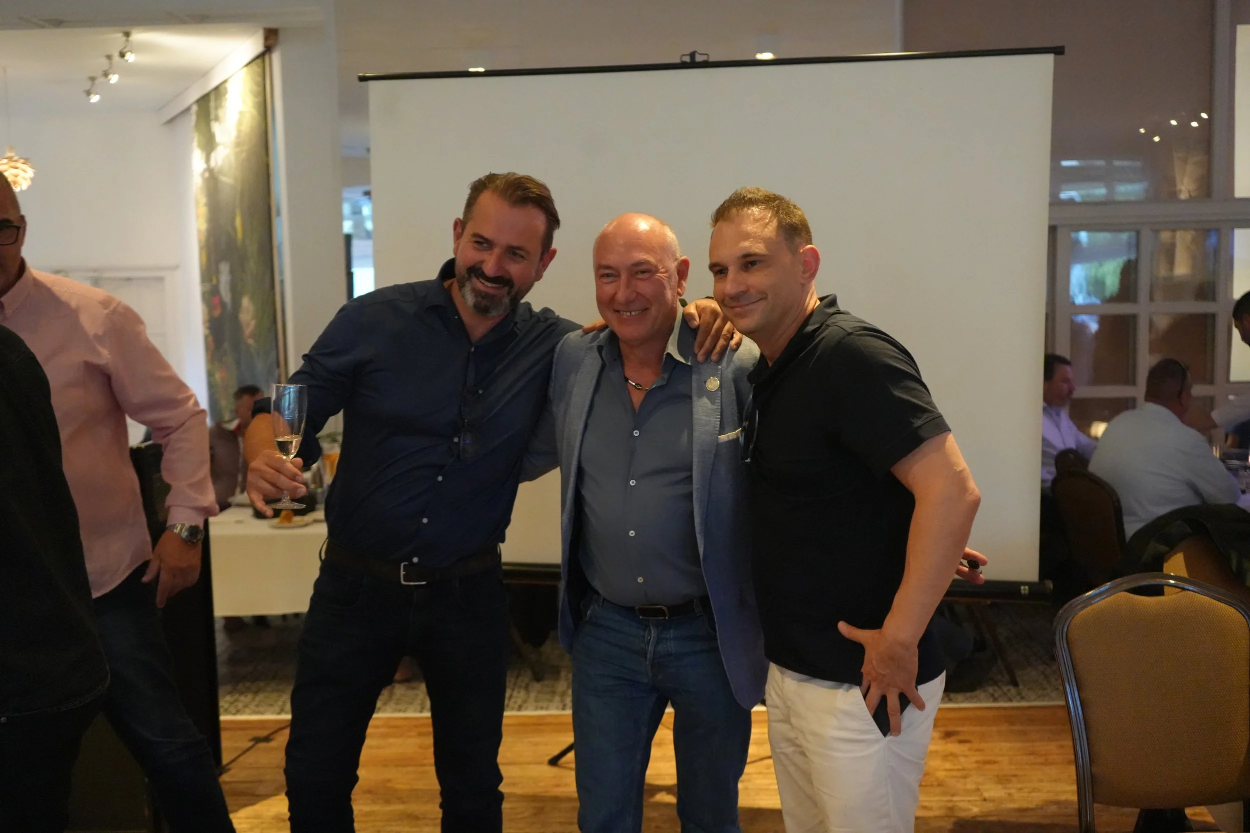 Three men standing together, smiling, in a social setting. One man holds a glass of champagne, and the other two have their arms around each other.