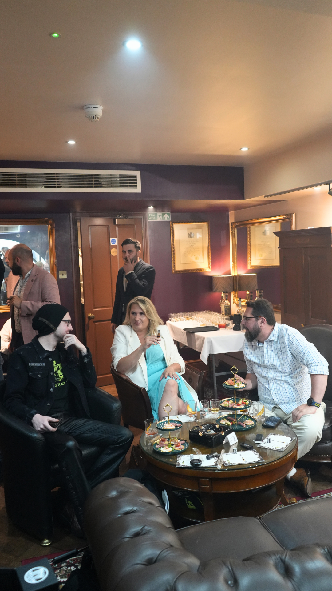 People socializing in a dimly lit lounge with a table of food and drinks.