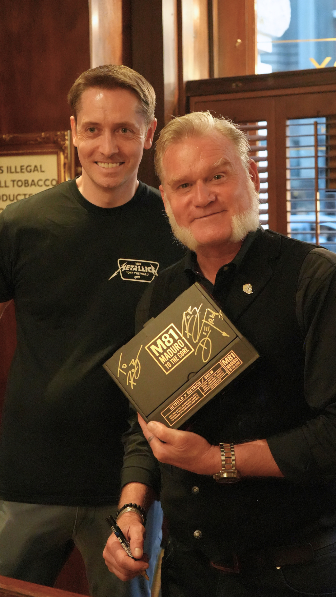 Two men smiling for a photo inside a restaurant or pub, with one man holding an autographed box with 'M81' and 'Maduro' written on it, and a signed message from a celebrity.