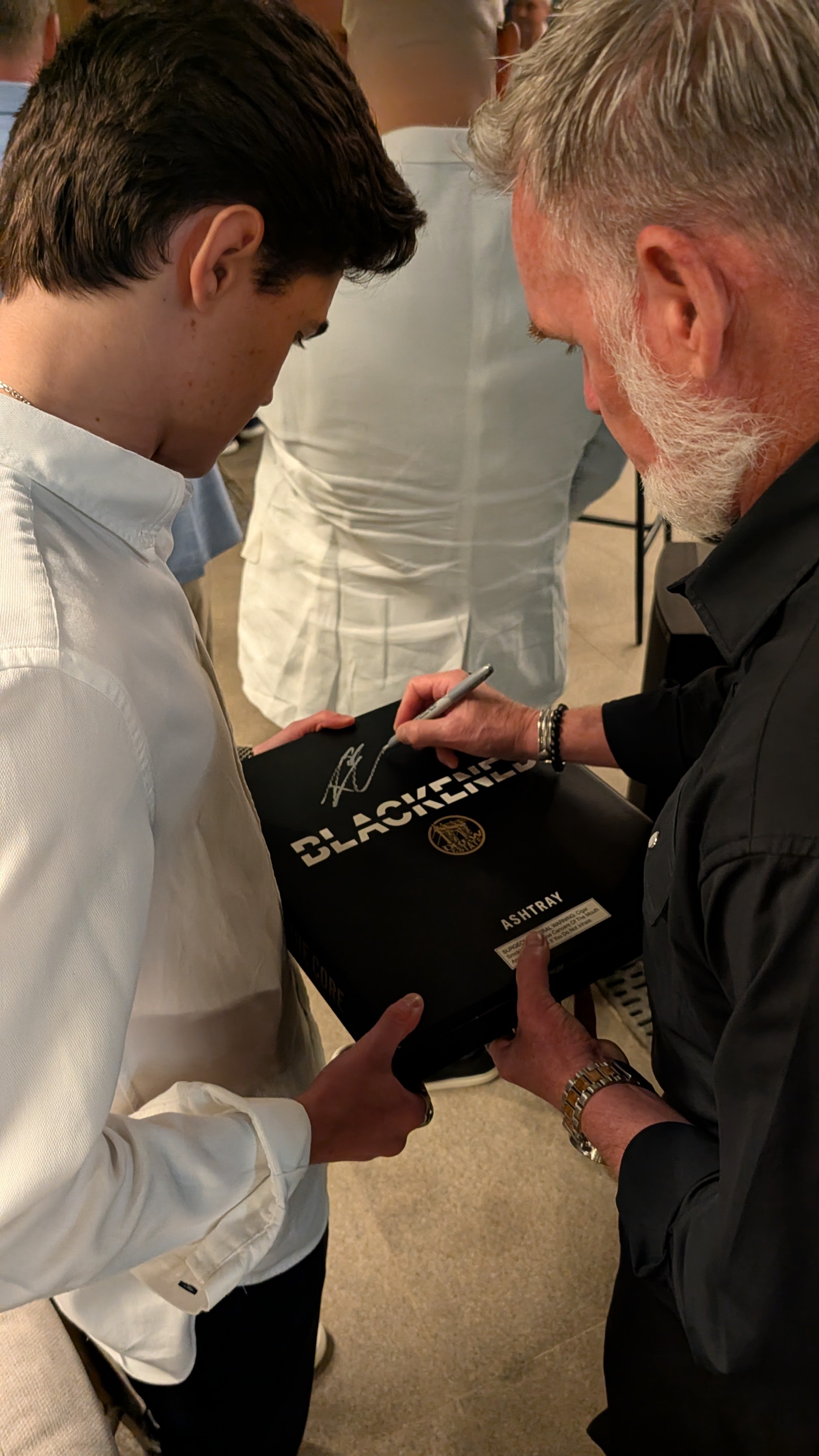 A man with gray hair and beard signing a black box or board labeled 'Blackened' held by a younger man with dark hair and light clothing.
