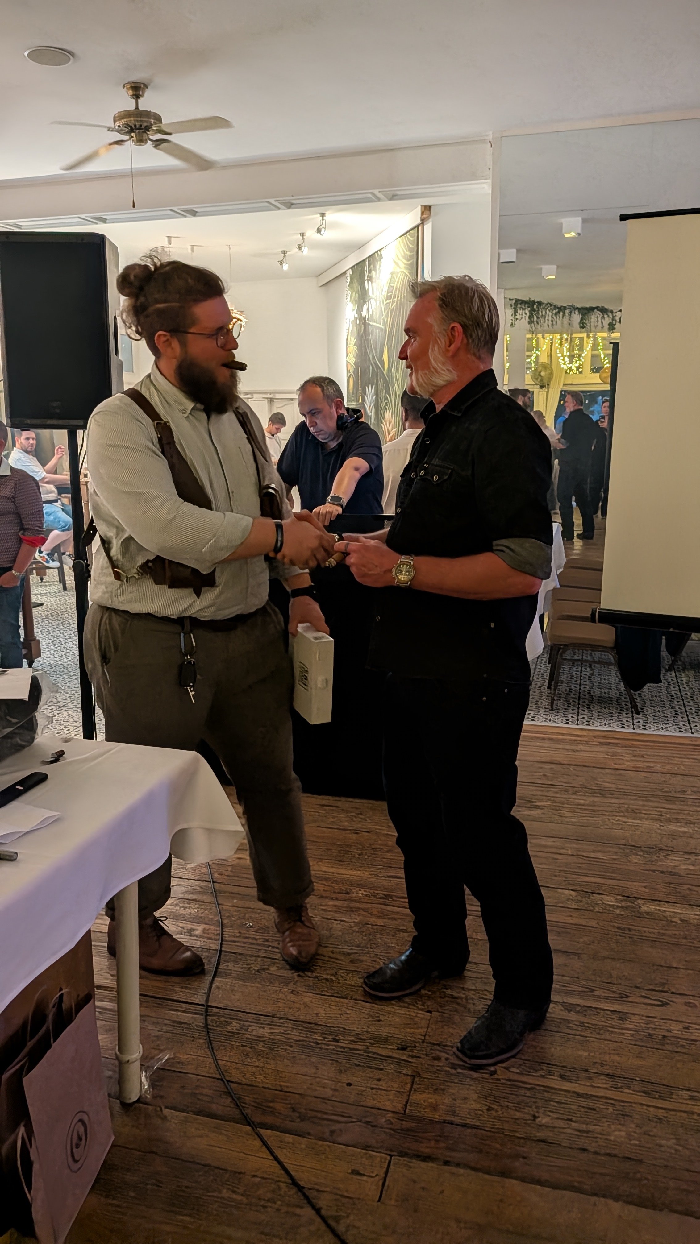 Two men shaking hands at an indoor event, one with a beard, glasses, and curly hair in a bun, and the other with gray hair and a beard, in a warmly lit room with people in the background.