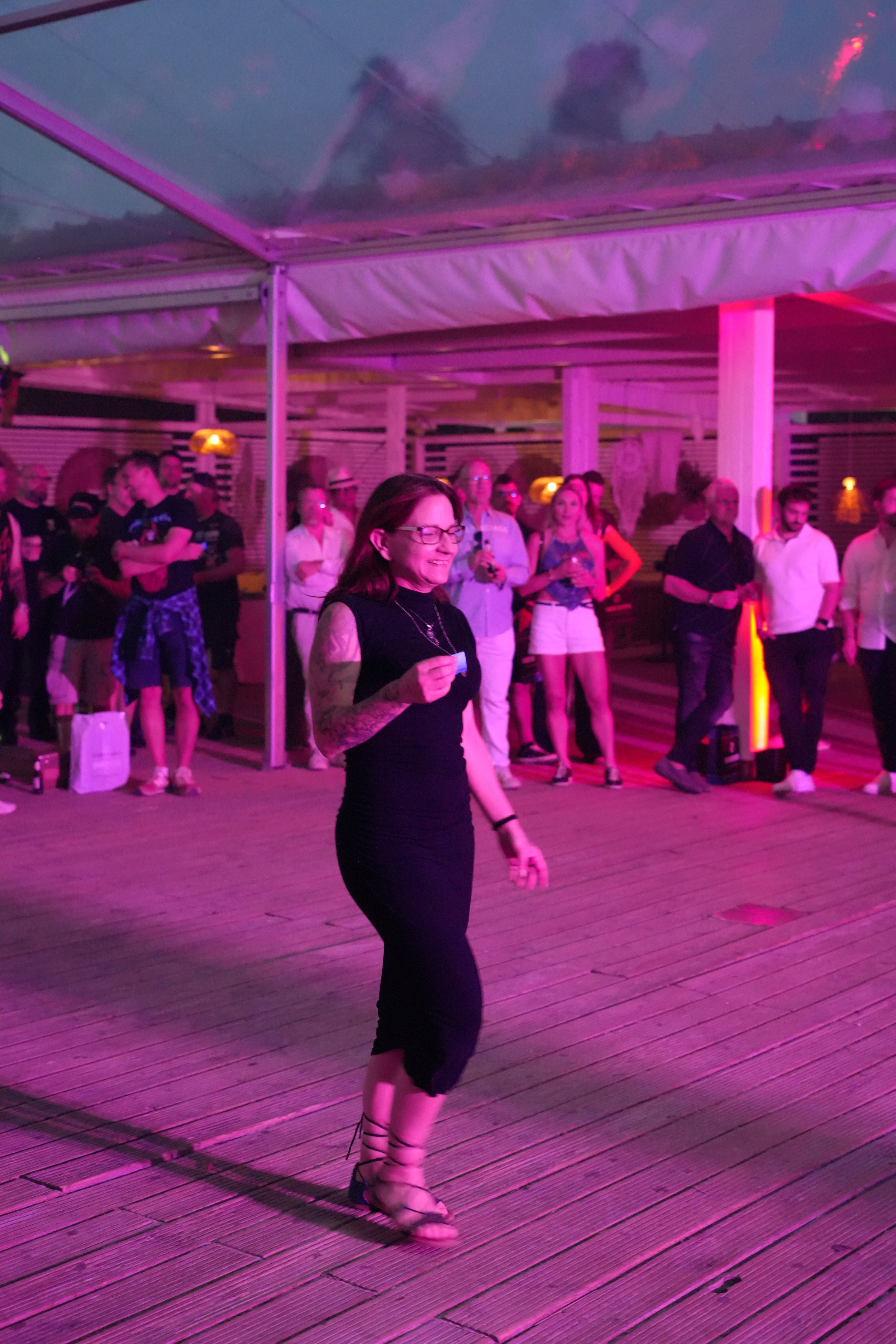 A woman with glasses and tattoos wearing black showing dance moves at a party or club with pink and purple lighting, surrounded by people watching.