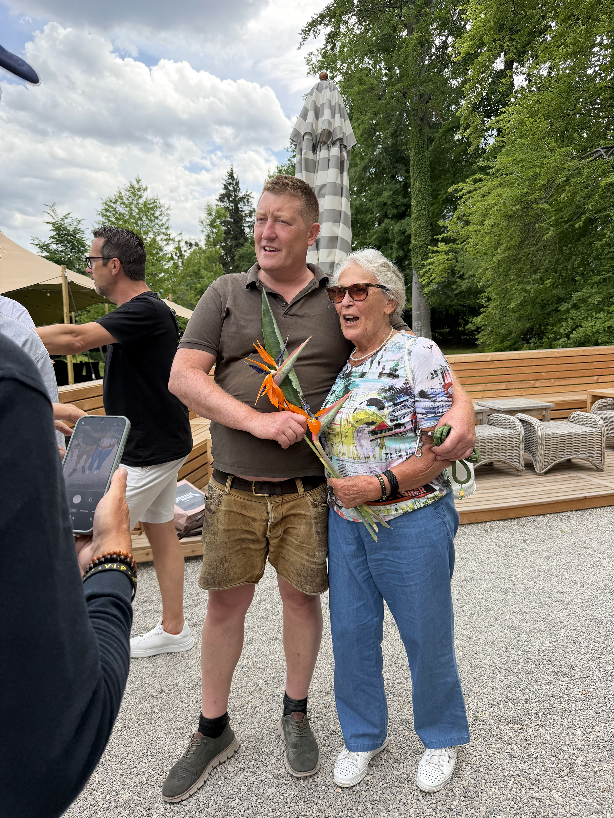 A man and an older woman are standing outdoors, smiling and holding a bouquet of flowers. They are surrounded by other people, with trees and a partly cloudy sky in the background.