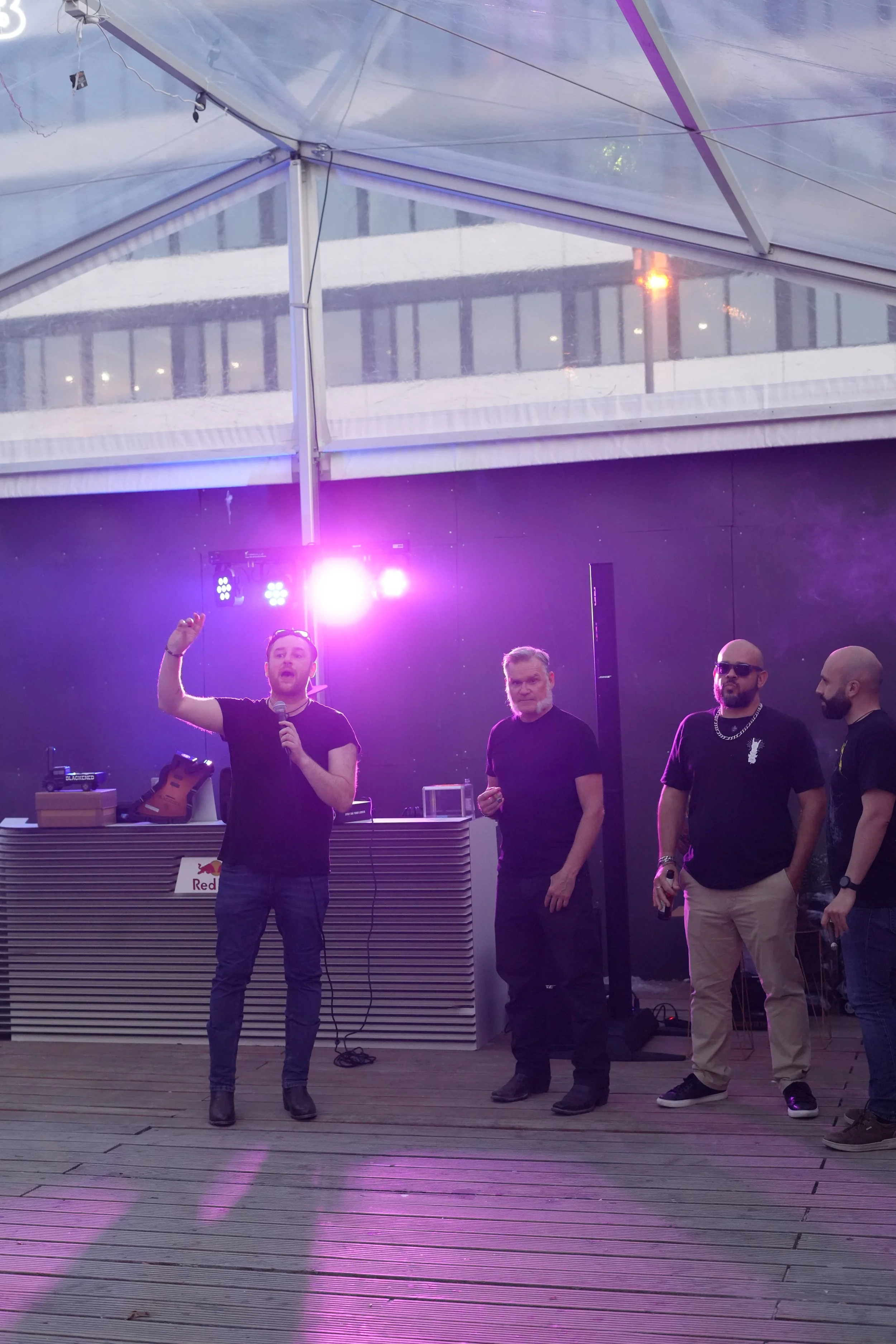 People standing and talking under a tent with purple lighting, with a DJ booth and equipment nearby. One person is holding a microphone, others are observing.