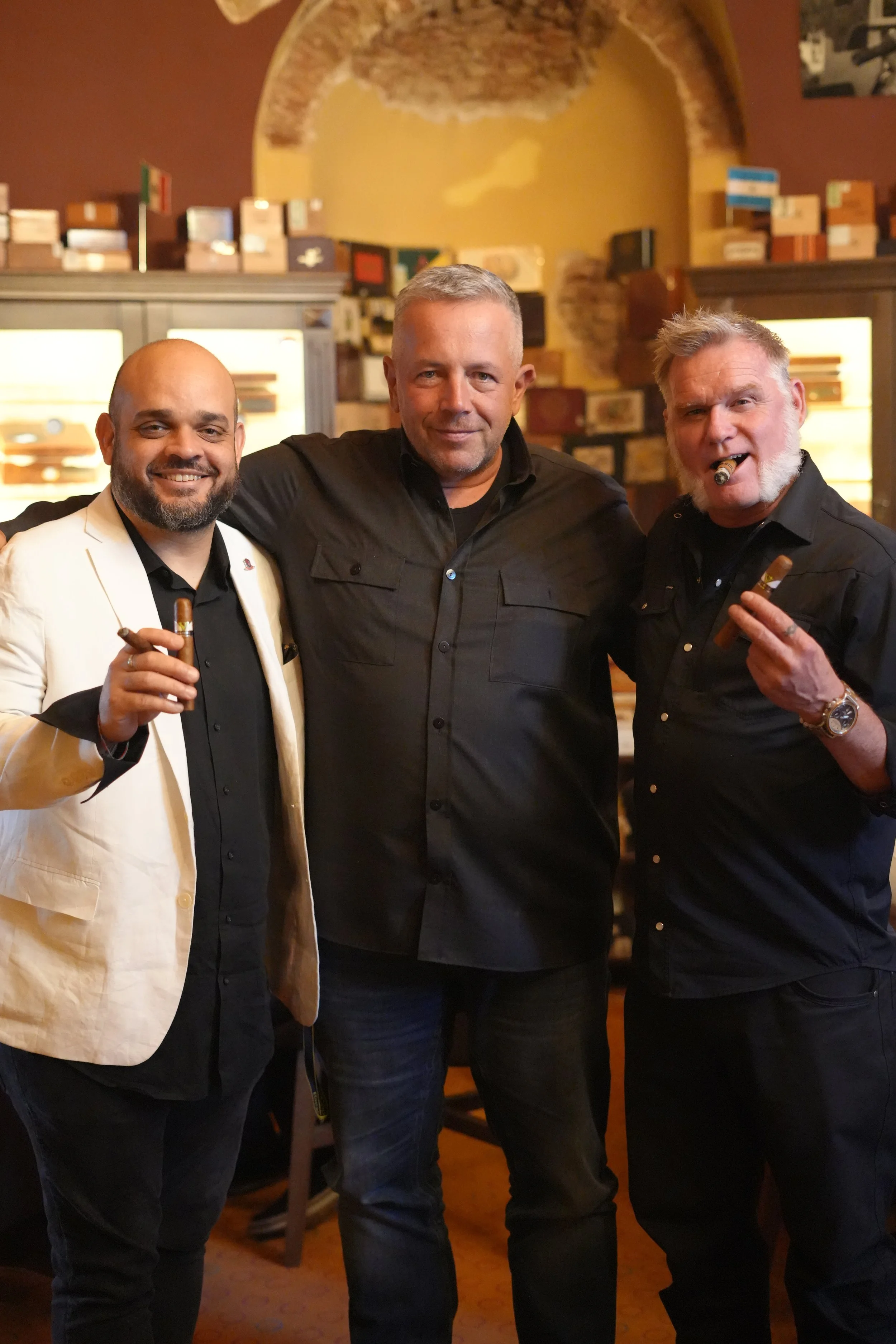 Three men standing together, two of them holding cigars, in a warm-lit indoor setting with shelves of boxes and a brick wall in the background.