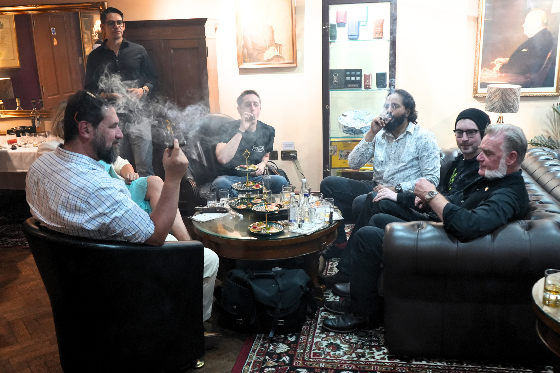Group of six men sitting in a living room, smoking and socializing around a table with appetizers and drinks, with one man standing in the background.