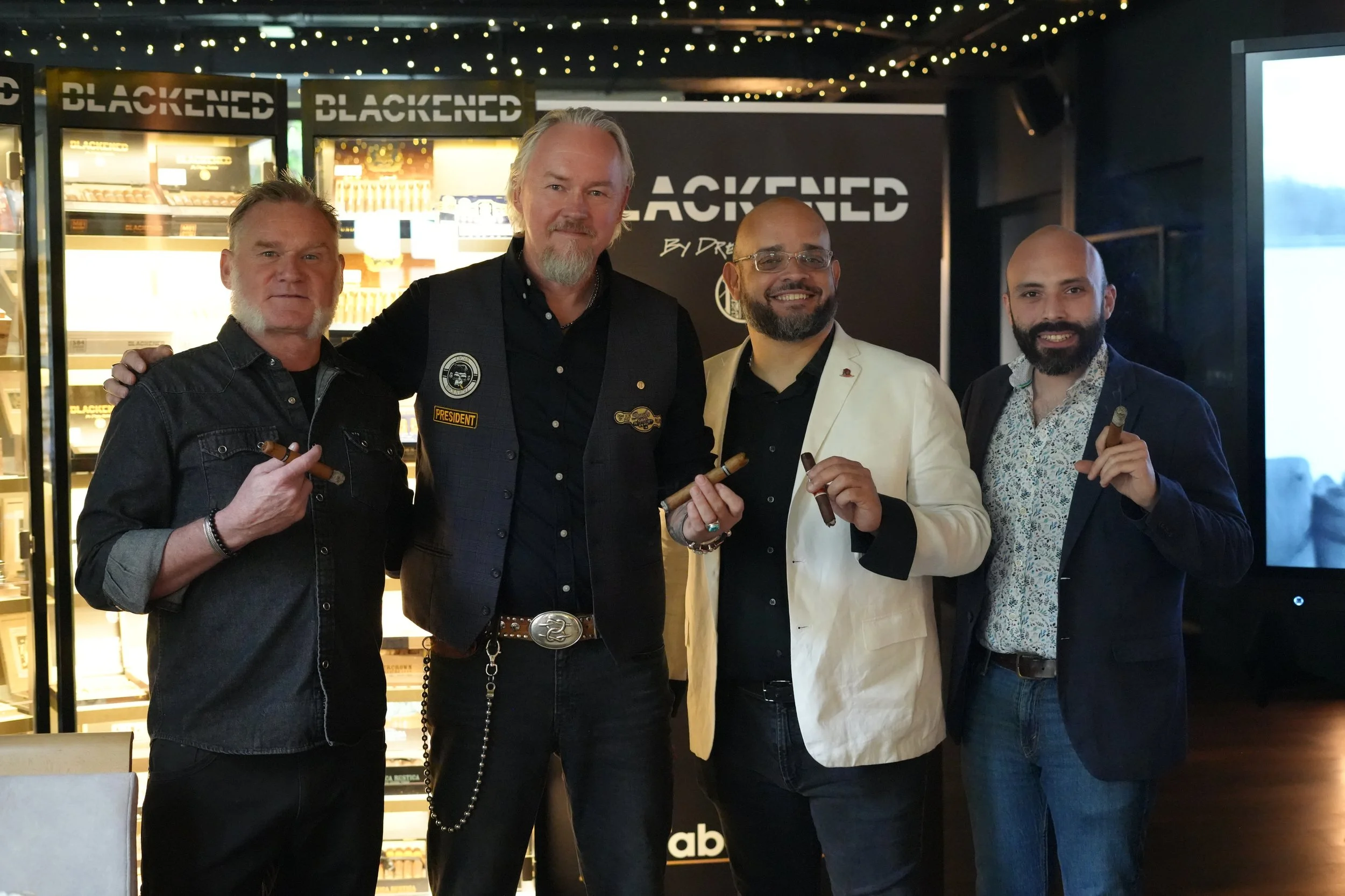 Four men standing together indoors, smiling and holding cigars, with a display of cigars and a black backdrop with white text that says 'BLACKENED' in the background.