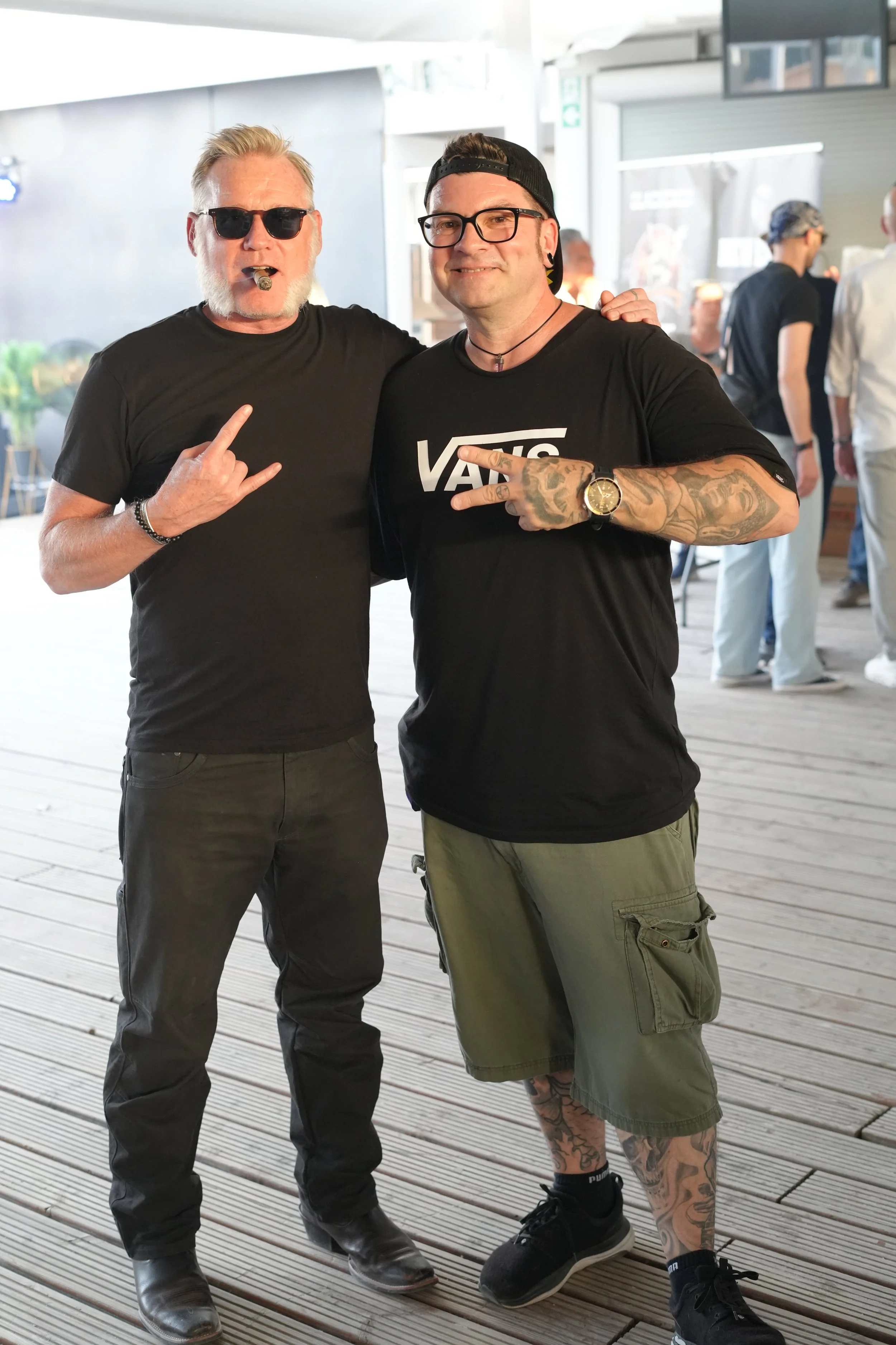 Two men standing indoors on a wooden floor, smiling and making gesture signs with their hands, with other people blurred in the background.