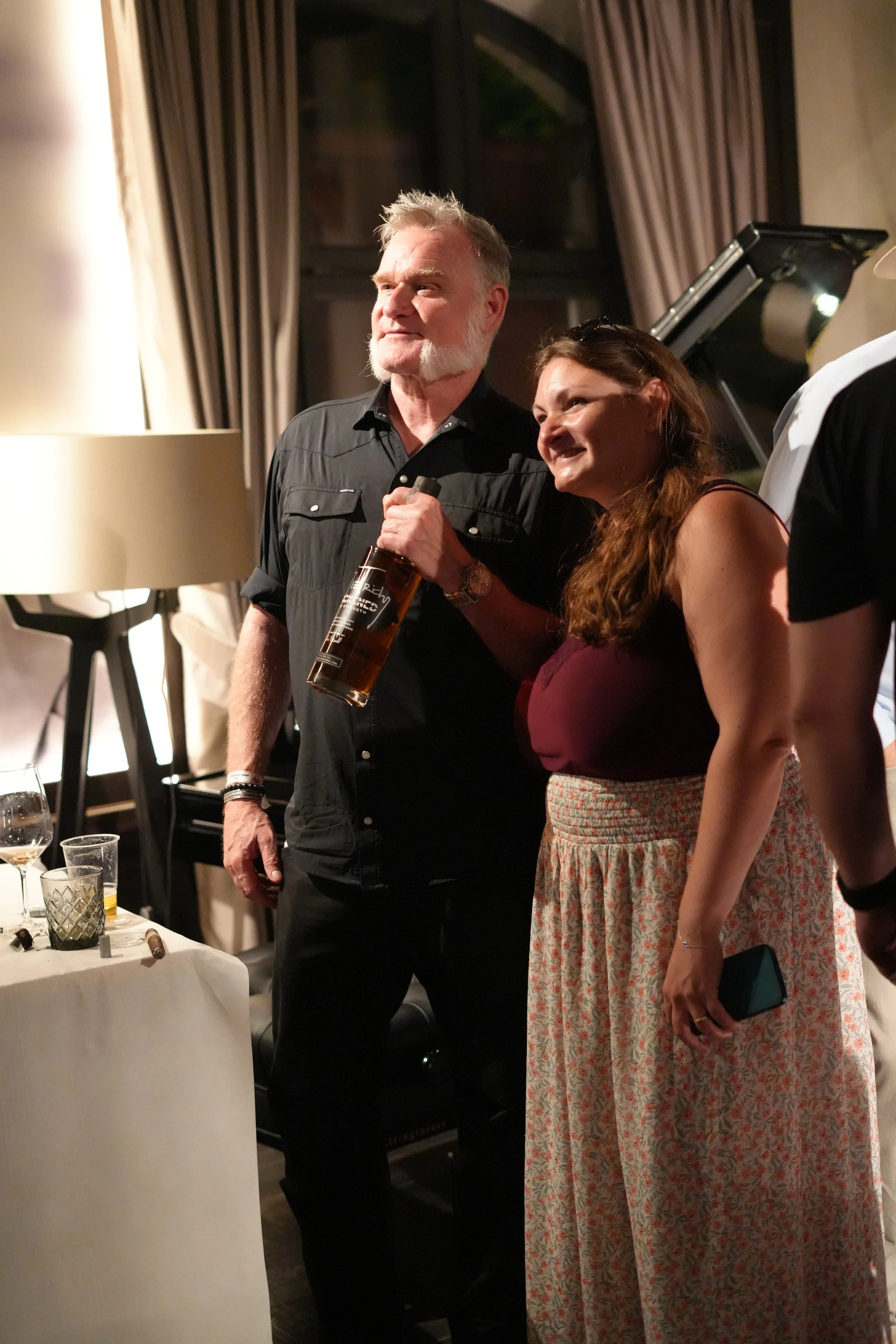 A man with gray hair and sideburns wearing a black shirt stands next to a woman with long brown hair dressed in a maroon top and floral skirt at a social gathering. The man holds a bottle of whiskey. There is a table with glasses and bottles in front