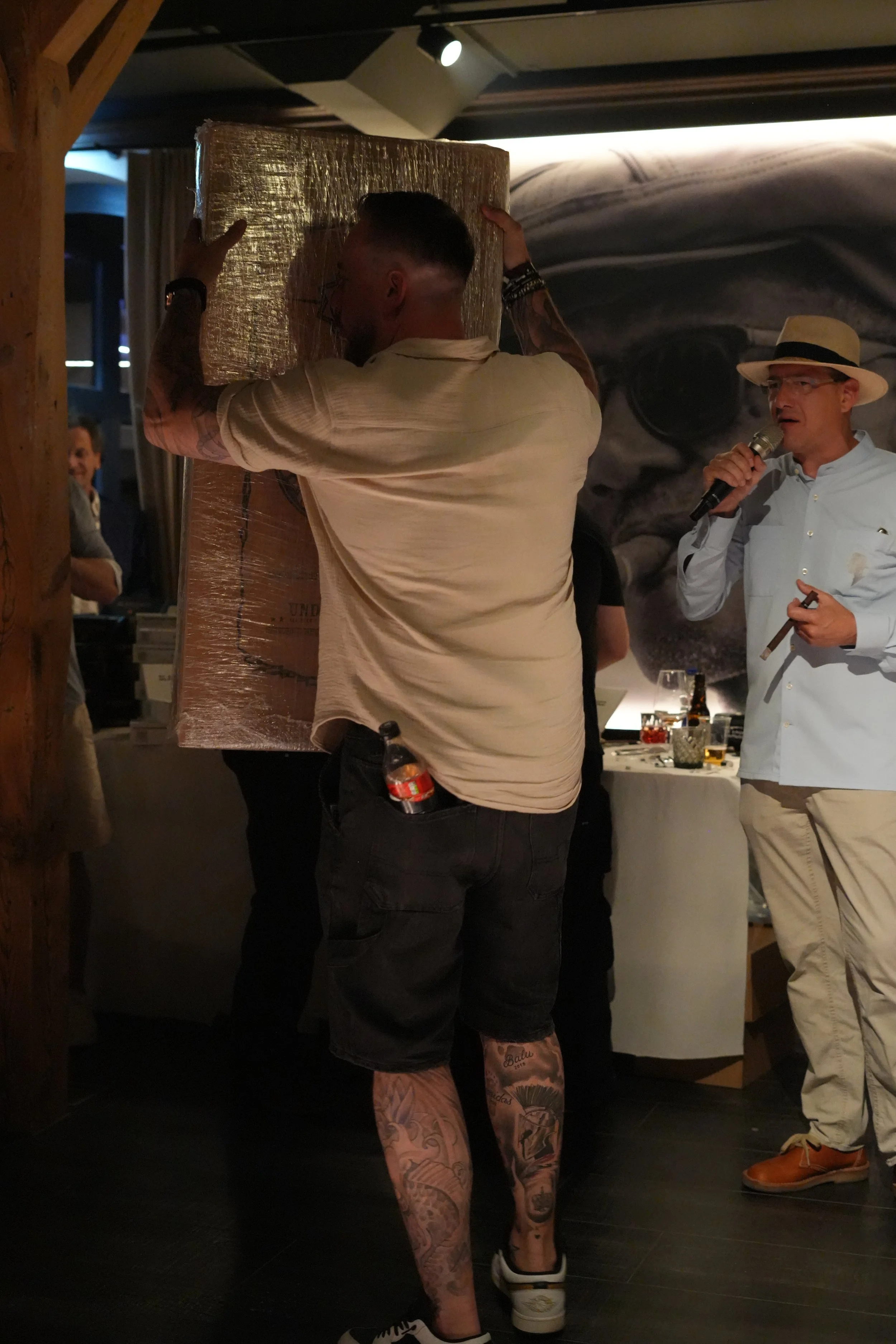 A man with tattoos carrying a wrapped pallet in a dimly lit room. Another man with a microphone, wearing a straw hat and glasses, stands nearby. A third person is visible in the background.