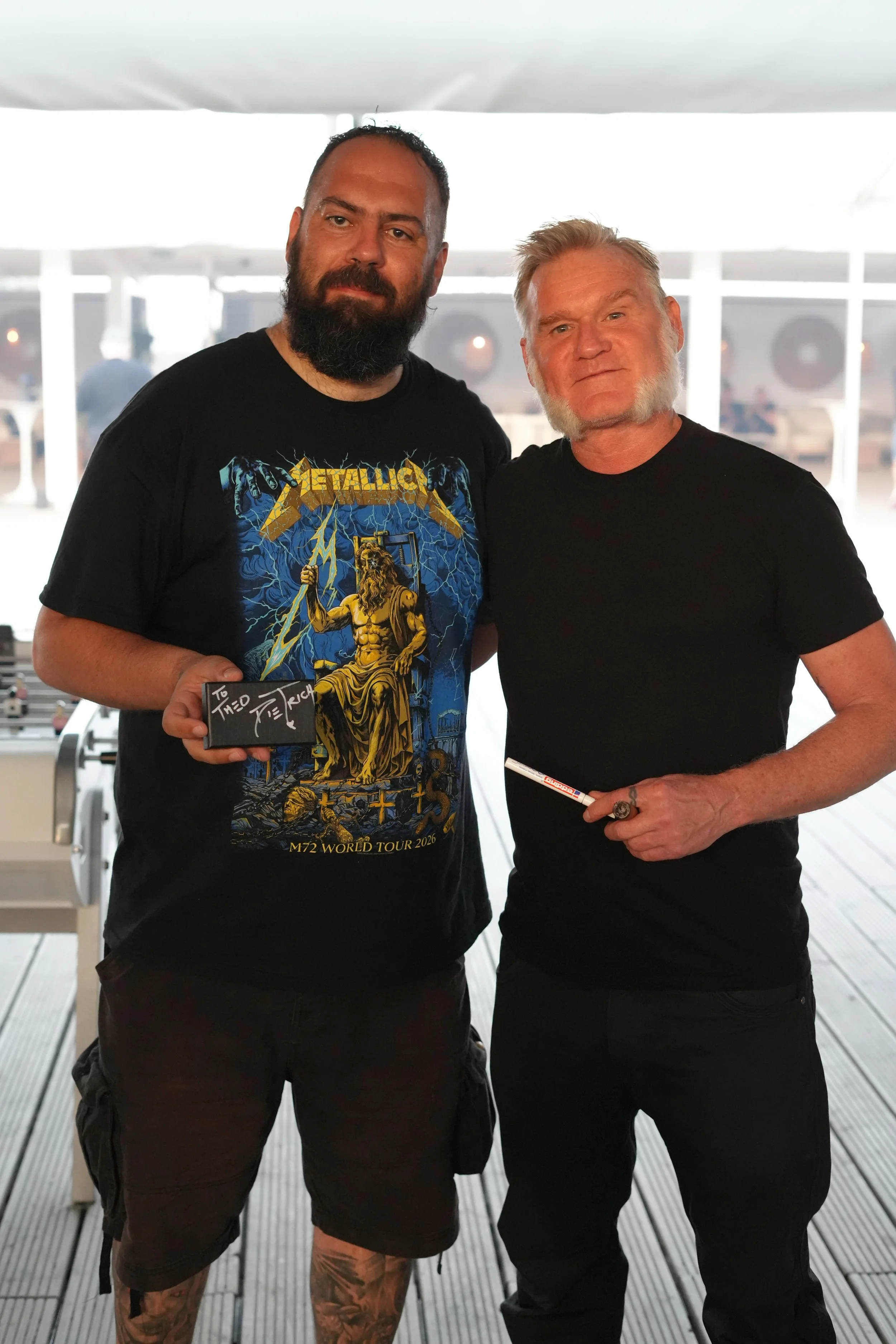 Two men standing together on a deck with a bright background, one wearing a Metallica t-shirt and the other in a black t-shirt, both holding items and posing for the photo.