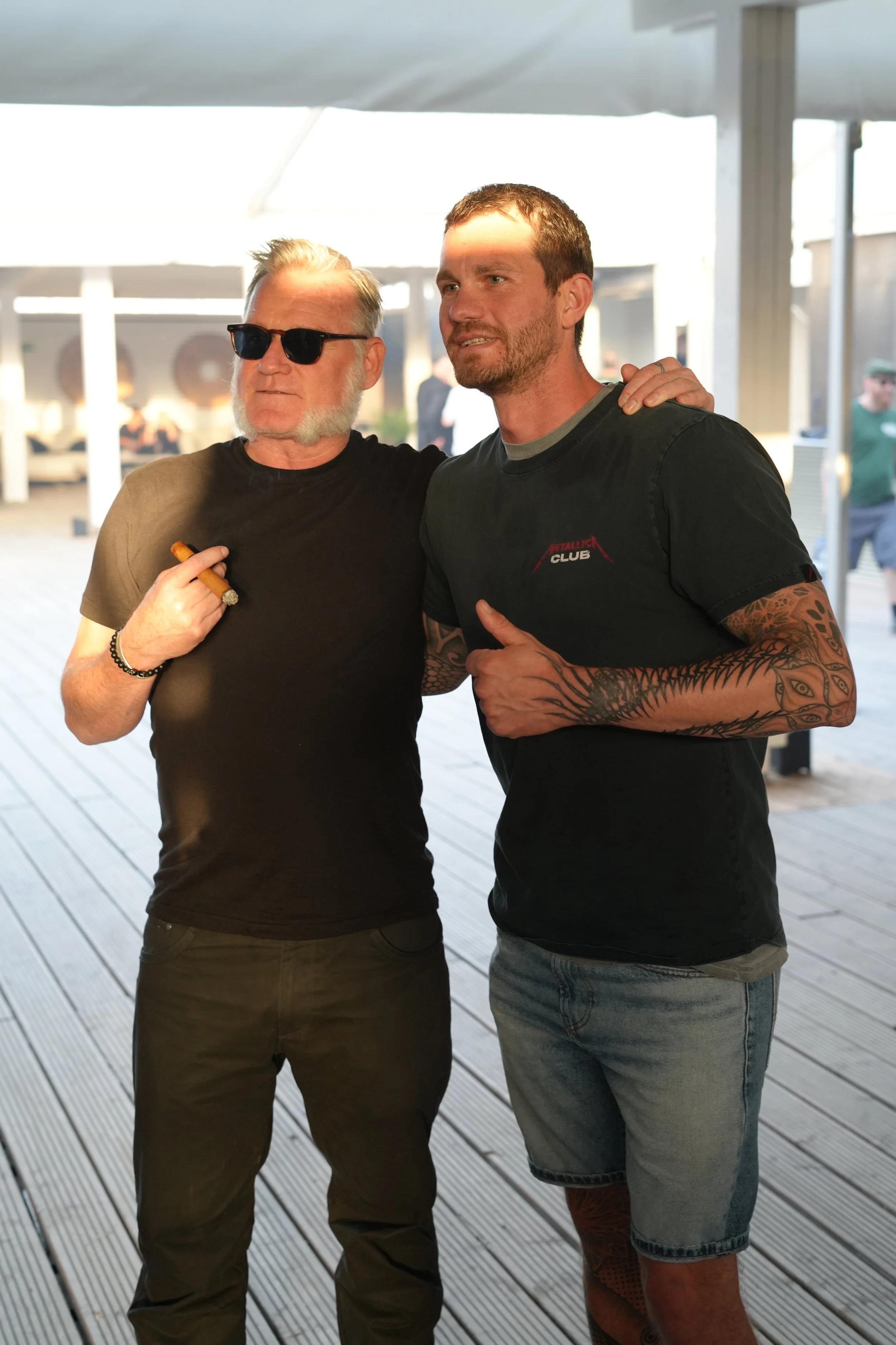 Two men posing together outdoors on a wooden deck, one with sunglasses and a cigar, the other with a tattooed arm, both smiling and relaxed.