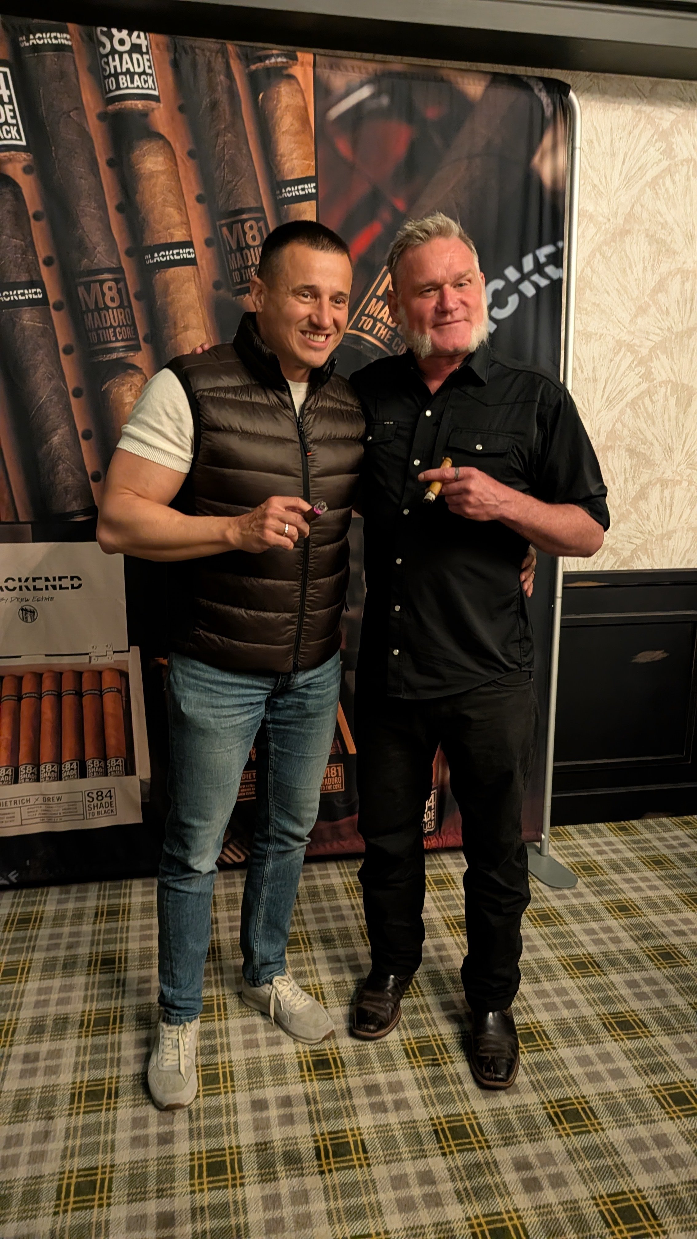 Two men standing together, smiling and holding cigars, in front of a backdrop with cigars and the text 'S84 Shade to Black' and 'M81 Maduro to the Core'.