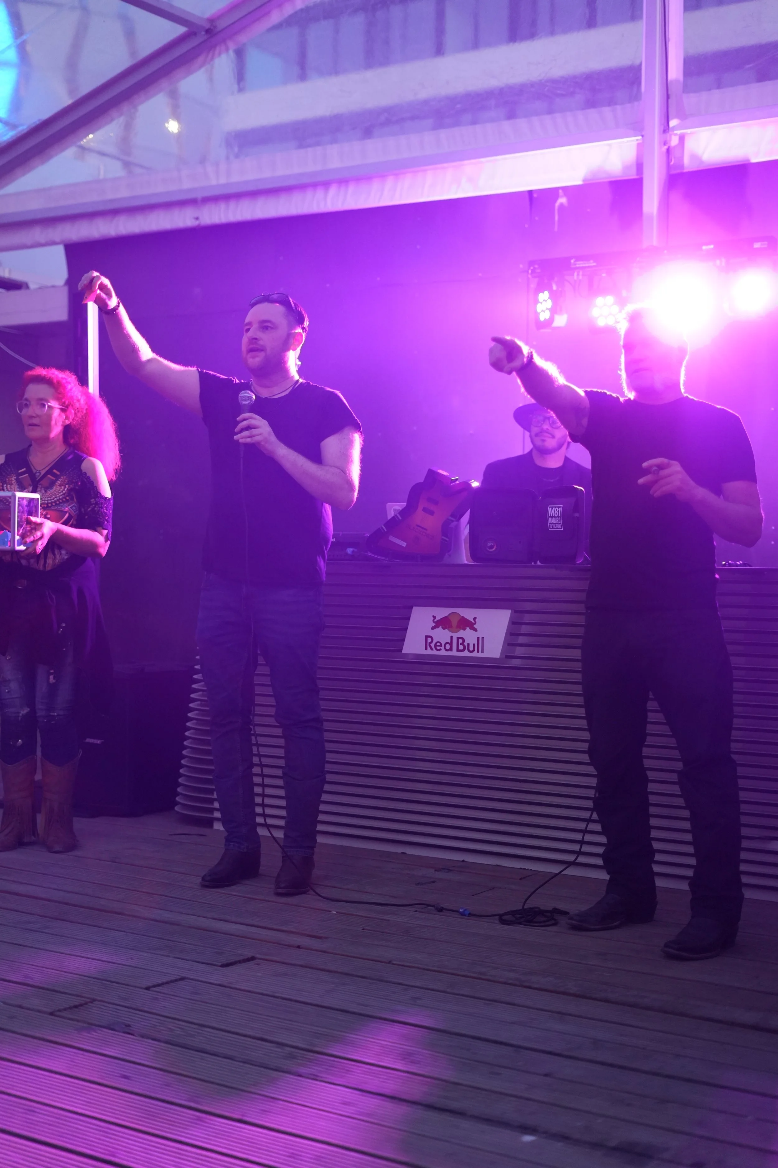Group of four people on a stage with purple lighting, one man holding a microphone and gesturing, others standing near DJ equipment with Red Bull logo, in an outdoor setting with a wooden floor.