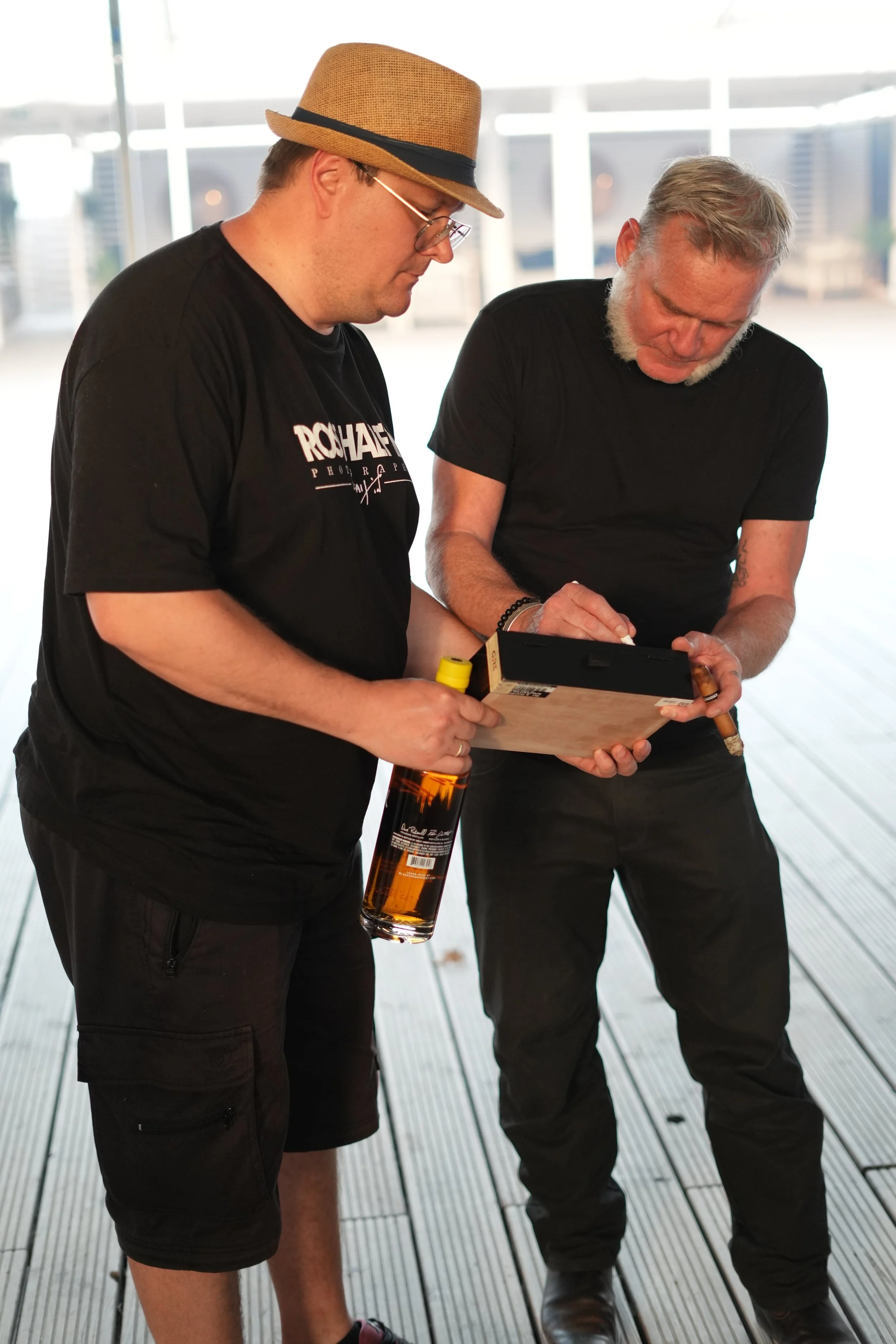 Two men standing outdoors on a wooden deck looking down at a small black object, with a cityscape visible through large windows in the background.