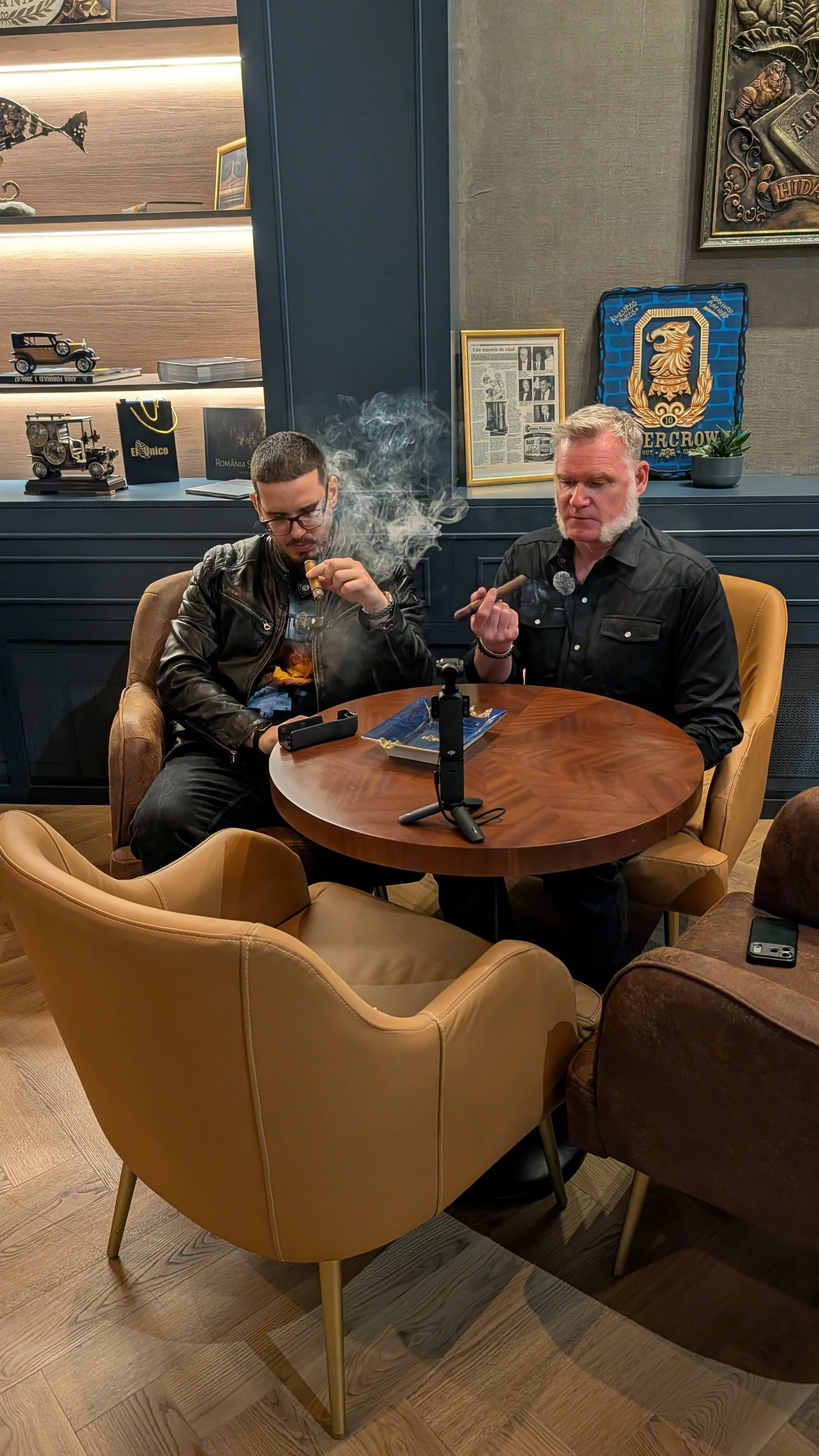 Two men sit at a round wooden table in a cozy, modern cafe, smoking cigars. Behind them are framed pictures and model cars on the shelf, with a blue wall and decorative artwork.
