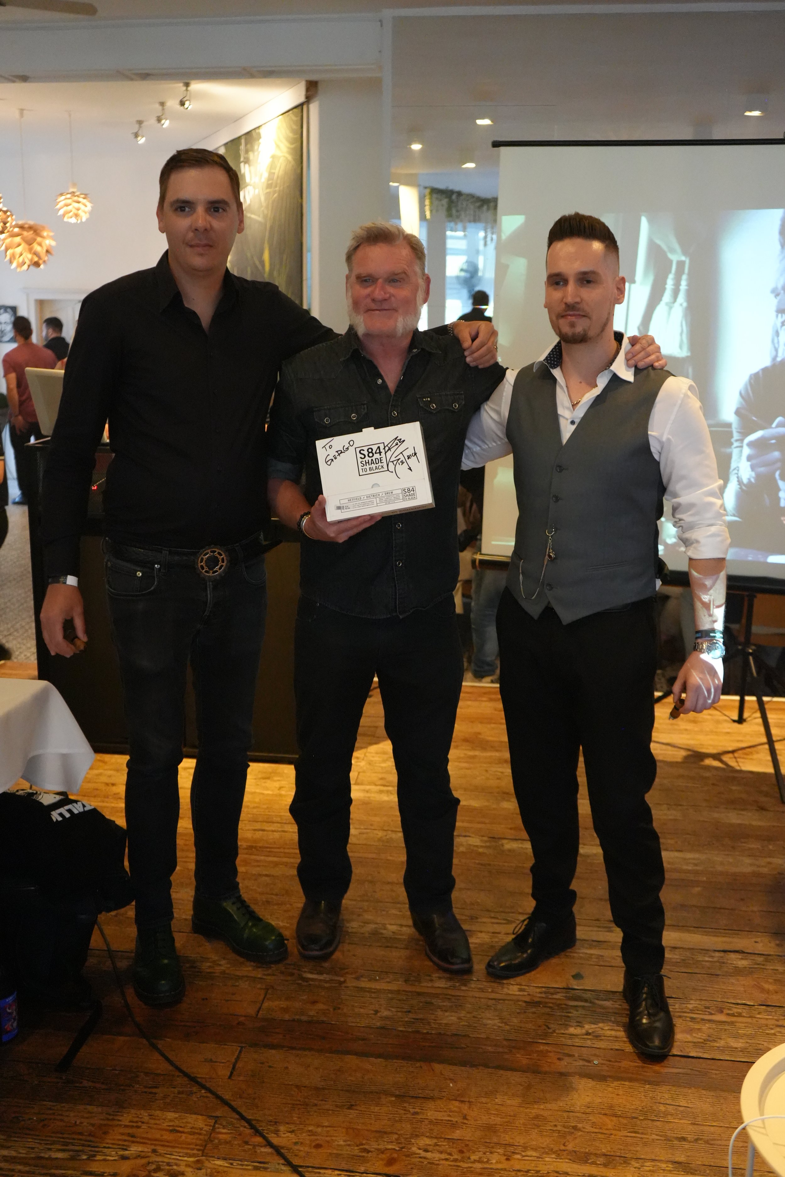 Three men standing side by side in an indoor setting, posing for a photo with one holding a box labeled 'S84 Shade to Black'.