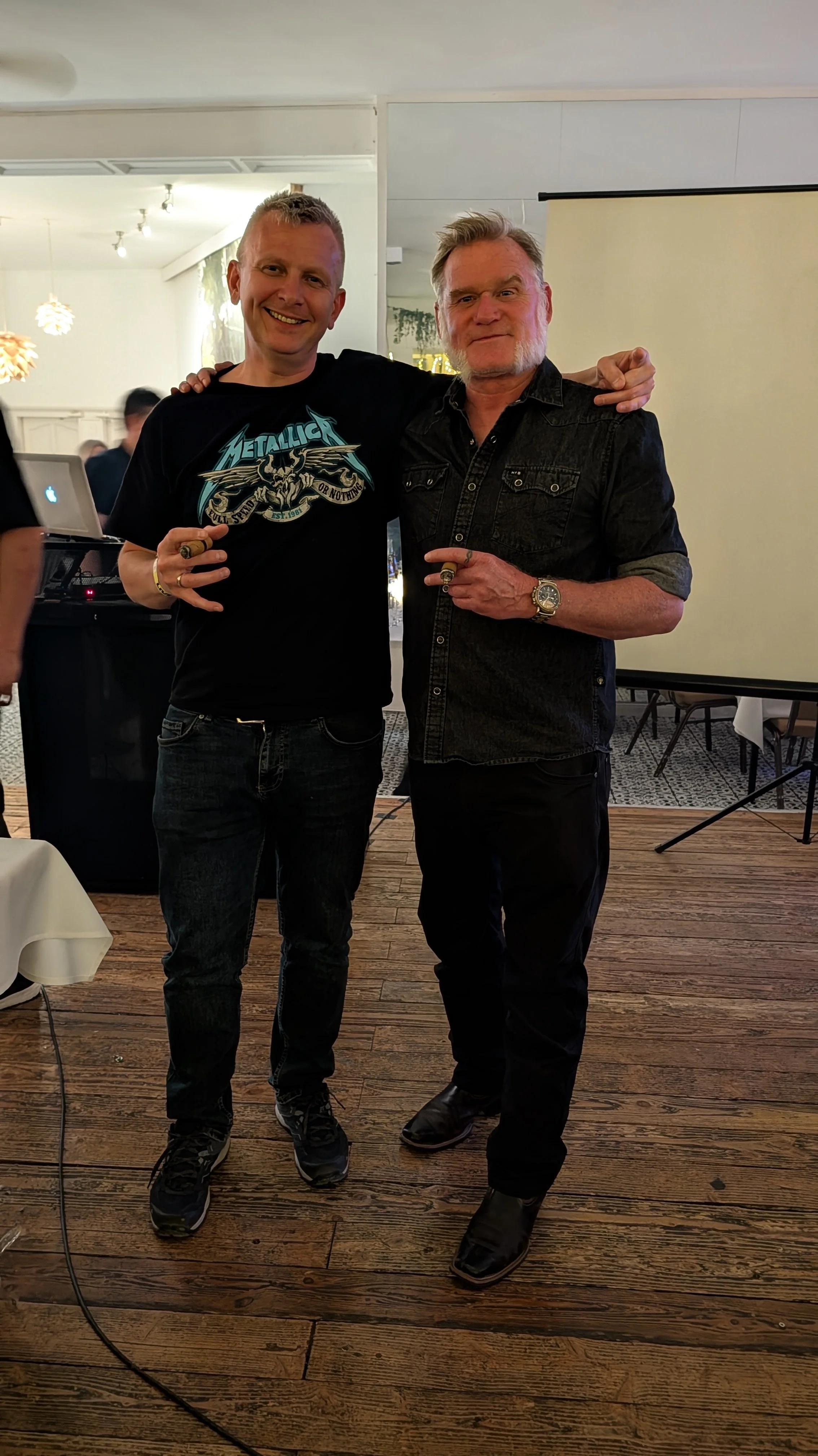Two men standing side by side in a room with wooden floors, smiling and posing for the camera. One is wearing a Metallica t-shirt and jeans, holding a ring, and the other is wearing a black denim shirt and dark pants, pointing at the man in the Metal