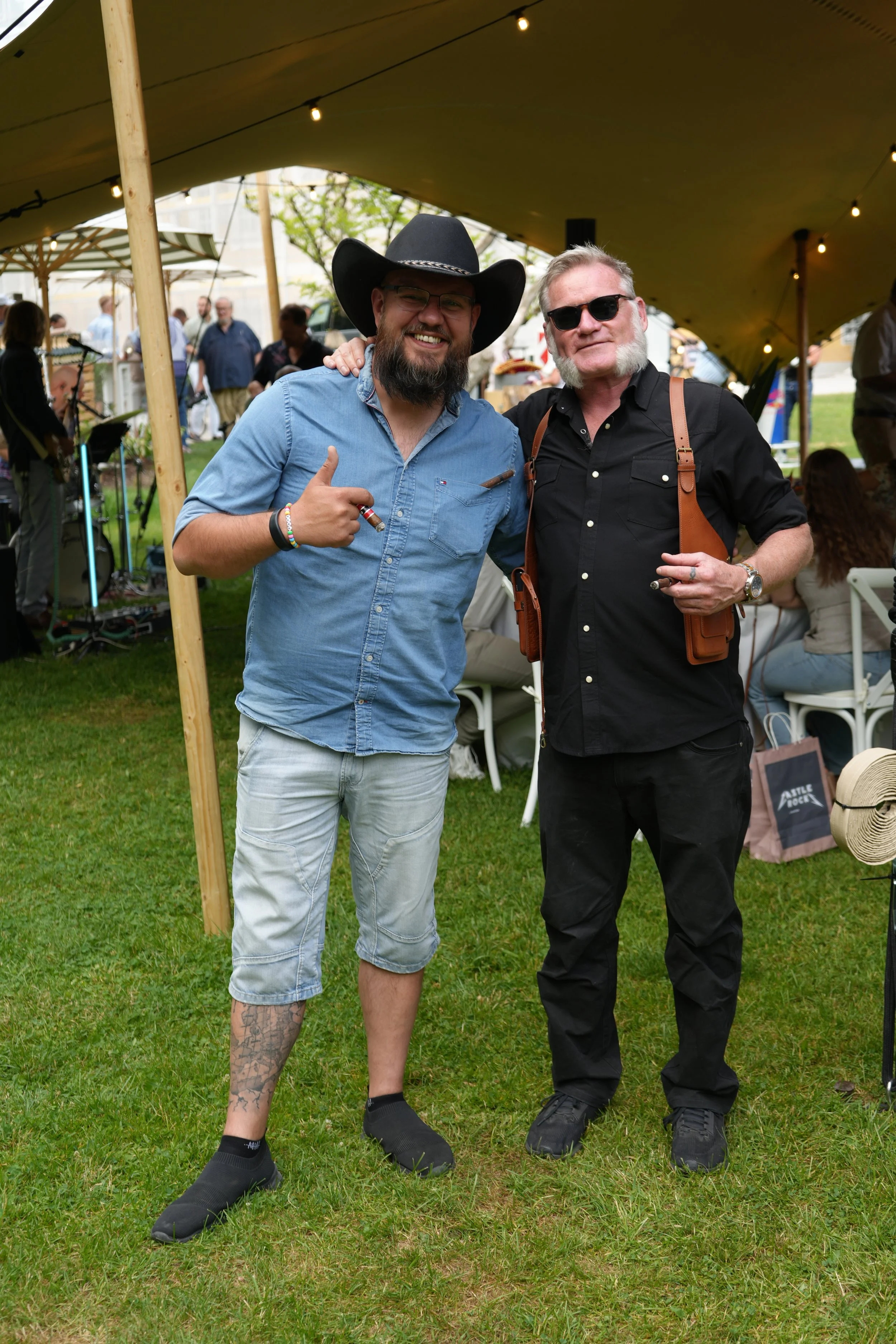 Two men standing close together outdoors at an event under a canopy, smiling at the camera. The man on the left wears a large black cowboy hat, glasses, a blue denim shirt, and light denim shorts, with tattoos visible on his legs. The man on the righ