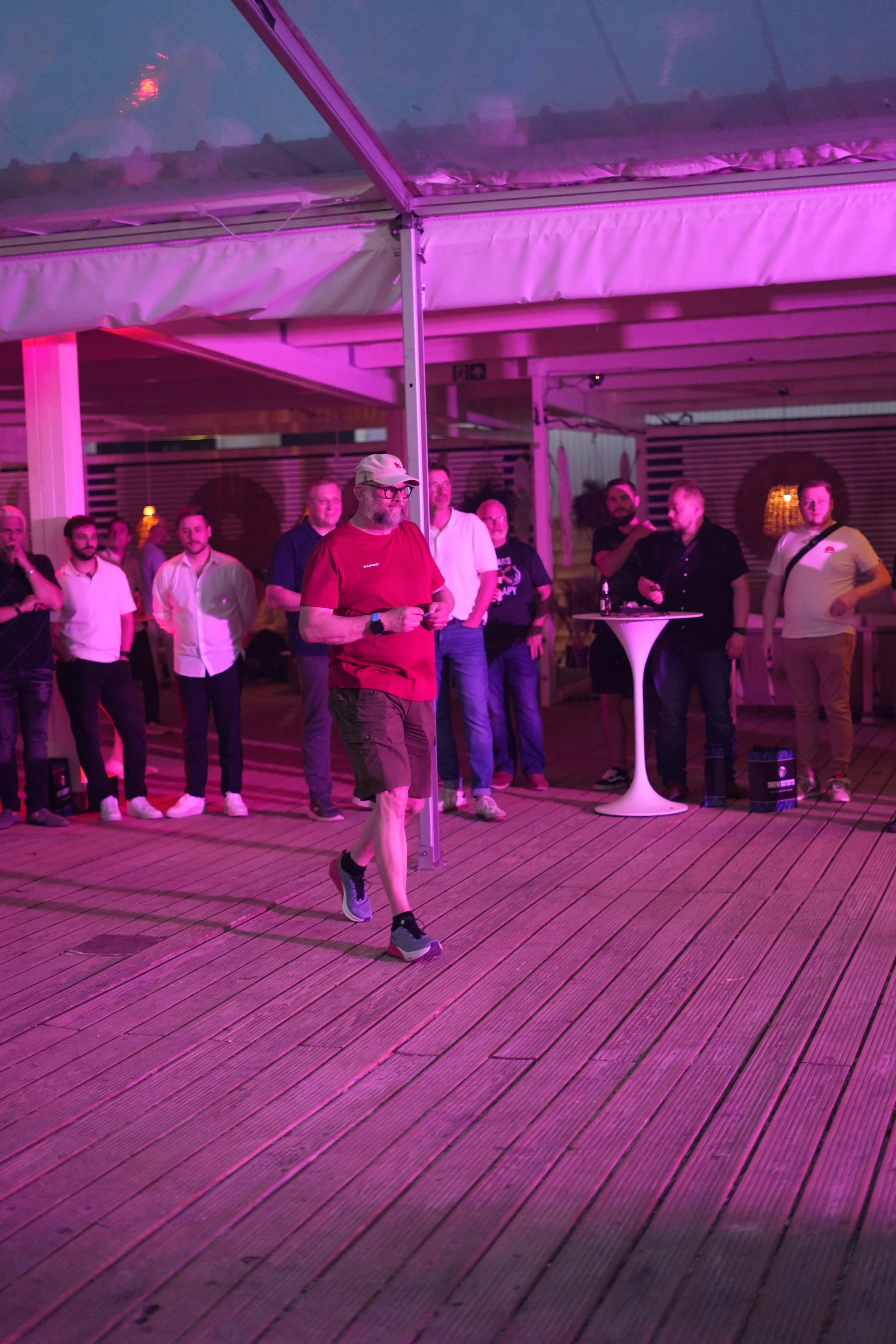 A man in a red shirt, shorts, and running shoes walking on a wooden deck under pink lighting. Several people are gathered in the background, some standing near a white cocktail table, with a purple-lit ceiling above.