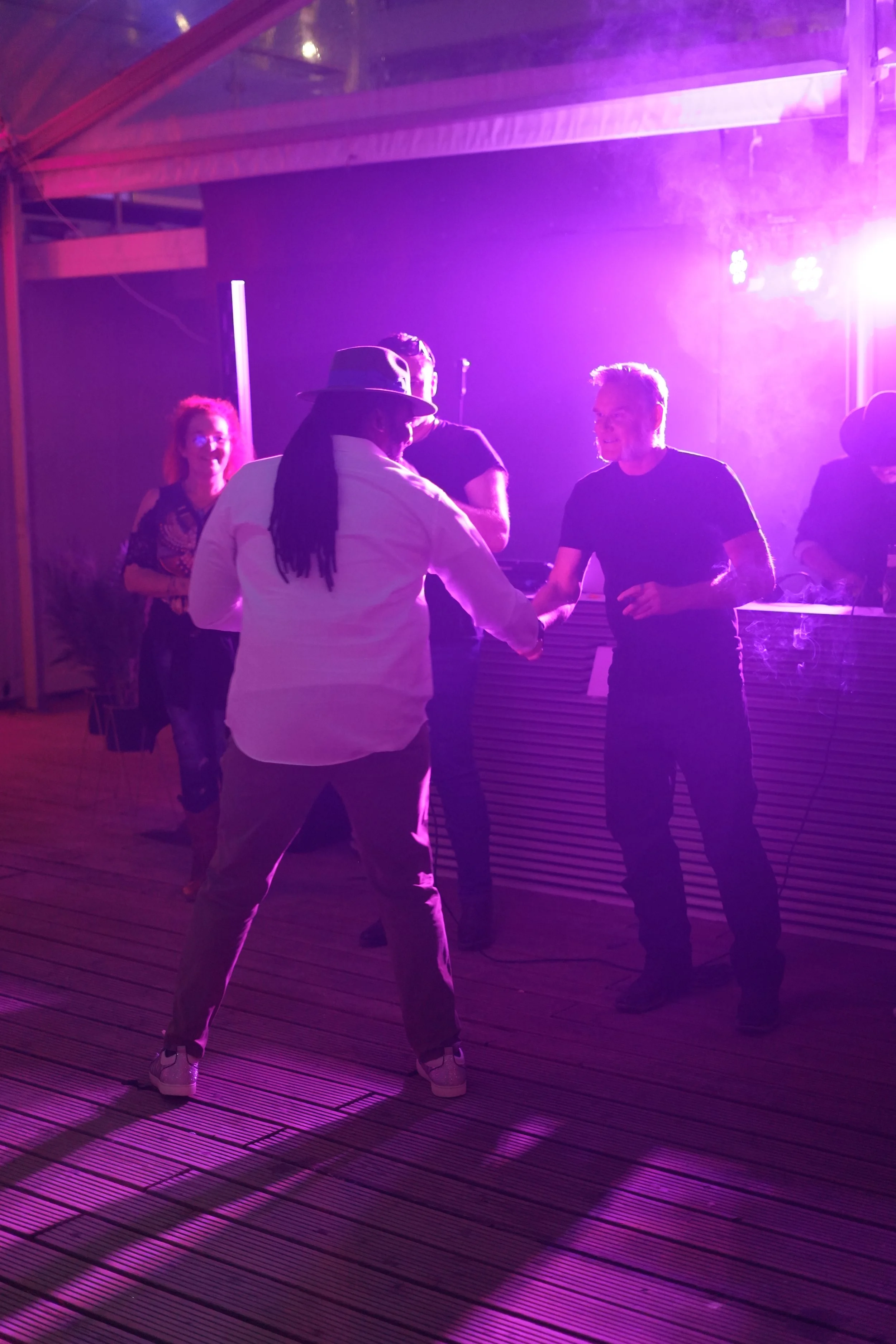 Group of people dancing and socializing at a party with purple lighting, on a wooden deck. One person in the foreground is wearing a white shirt and a hat, shaking hands with another person in a black shirt.