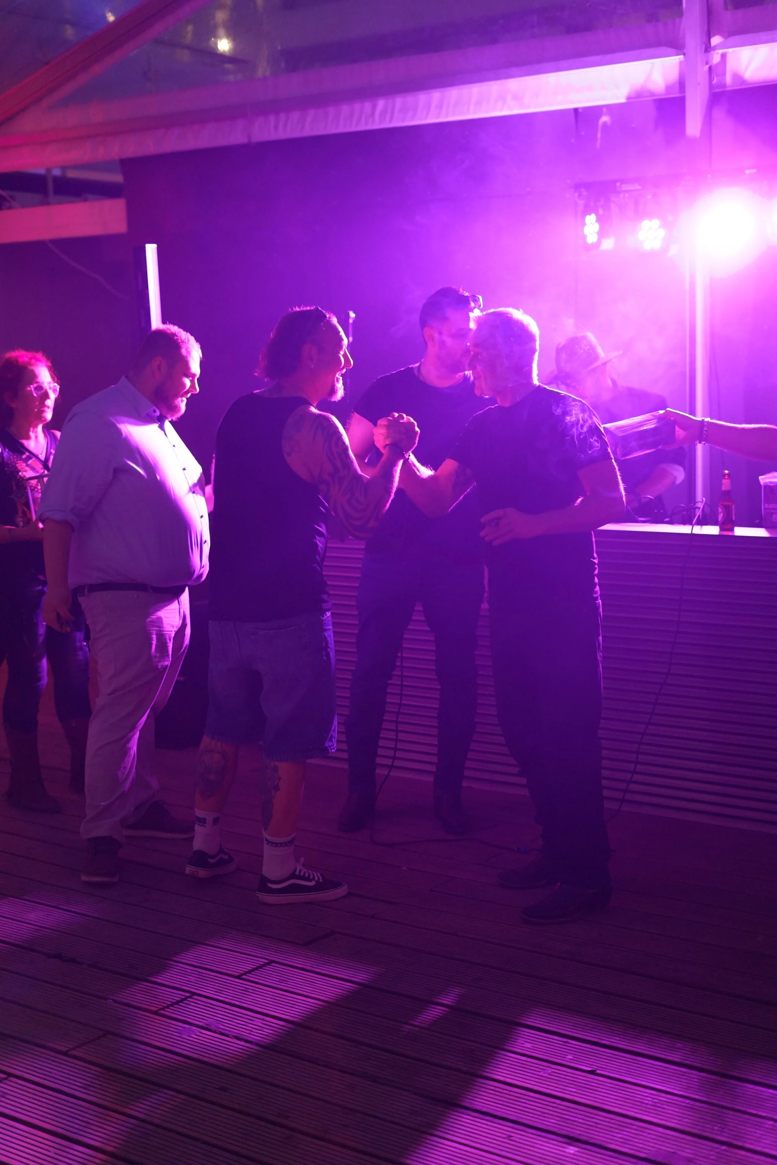 People socializing at a party with purple lighting, standing on a wooden deck, some shaking hands, others watching.
