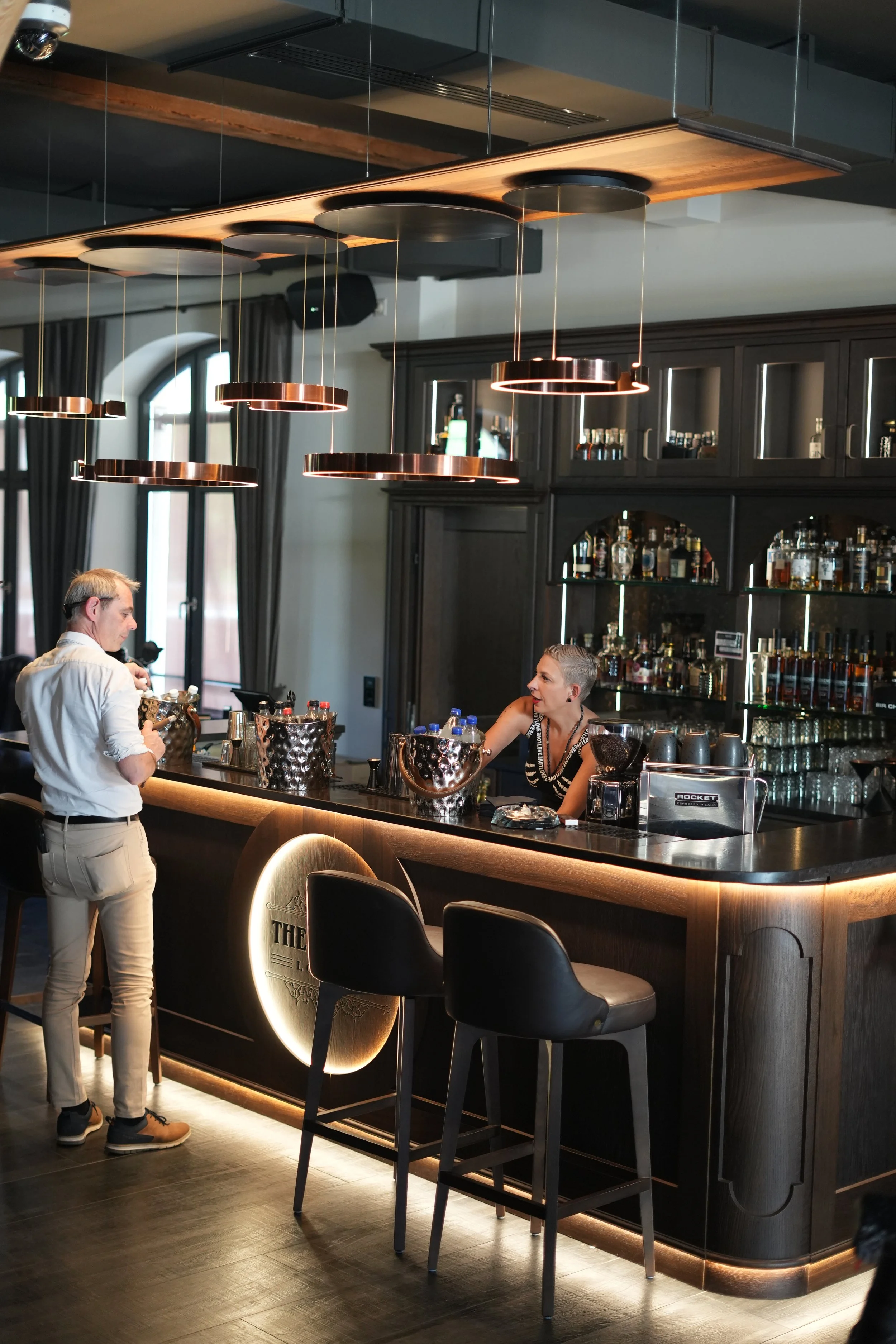 A bar scene with a woman talking to a bartender and a man ordering drinks at the counter. The bar has dark wood finishes, modern lighting fixtures, and shelves stocked with bottles in the background.