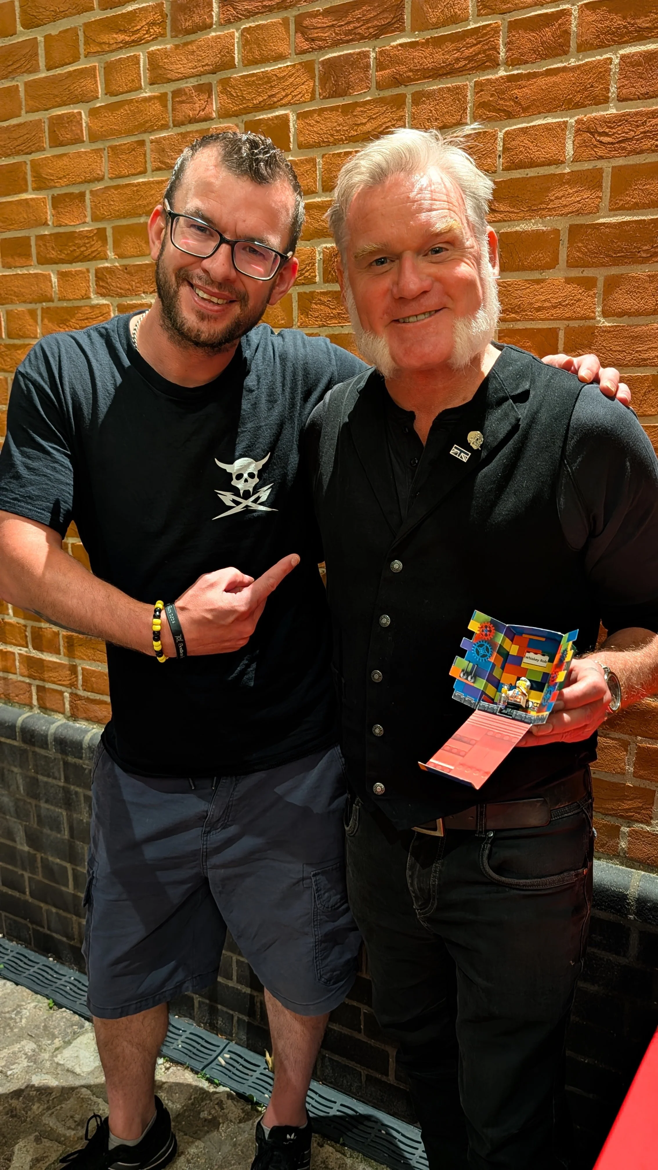 Two men standing in front of a brick wall. The man on the left is pointing at the man on the right, who is holding a colorful Lego artwork.