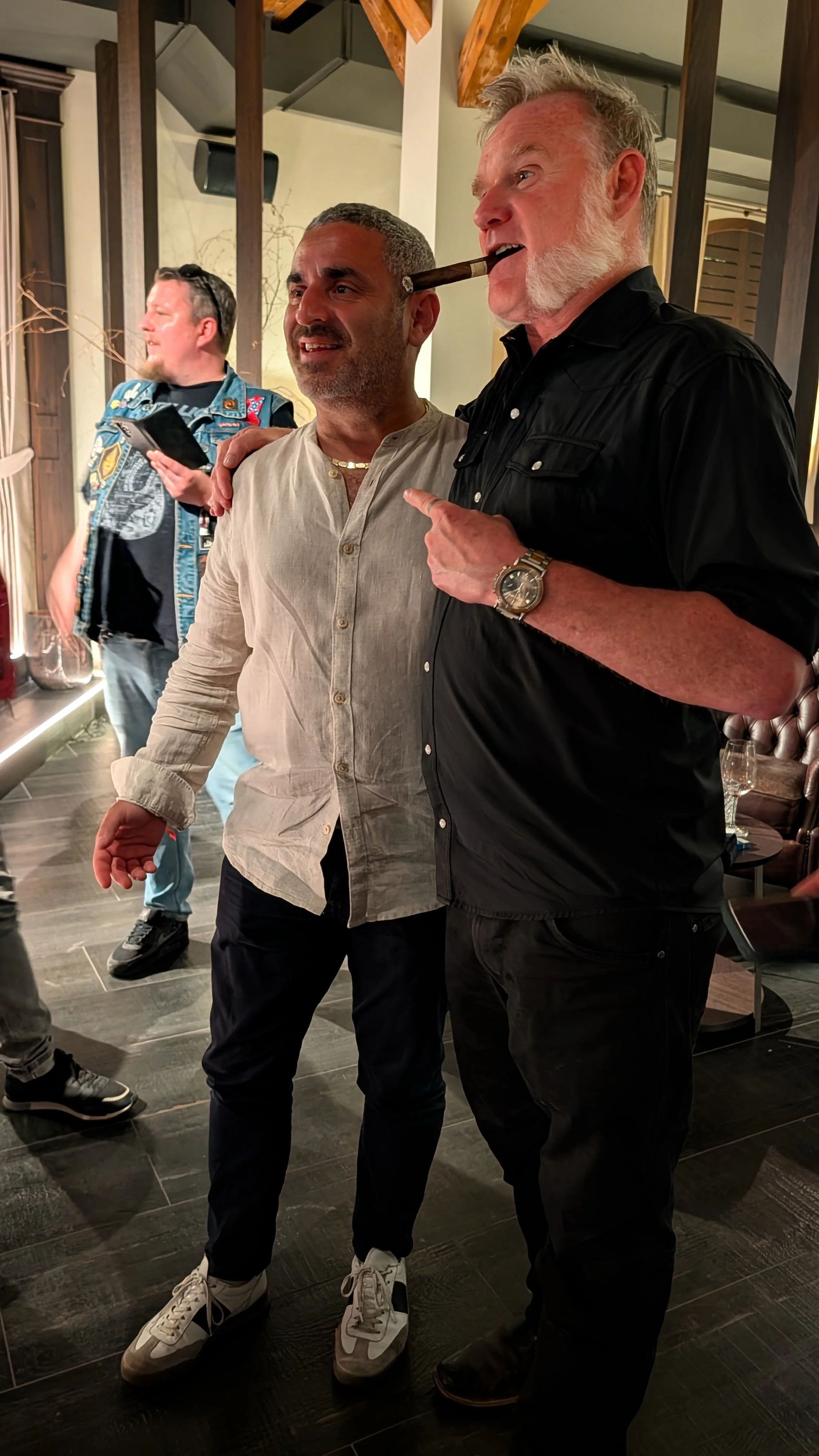 Two men standing close together in a social gathering, one with a cigar in his mouth pointing at the other. Both appear relaxed and are smiling. In the background, another man is looking at his phone.