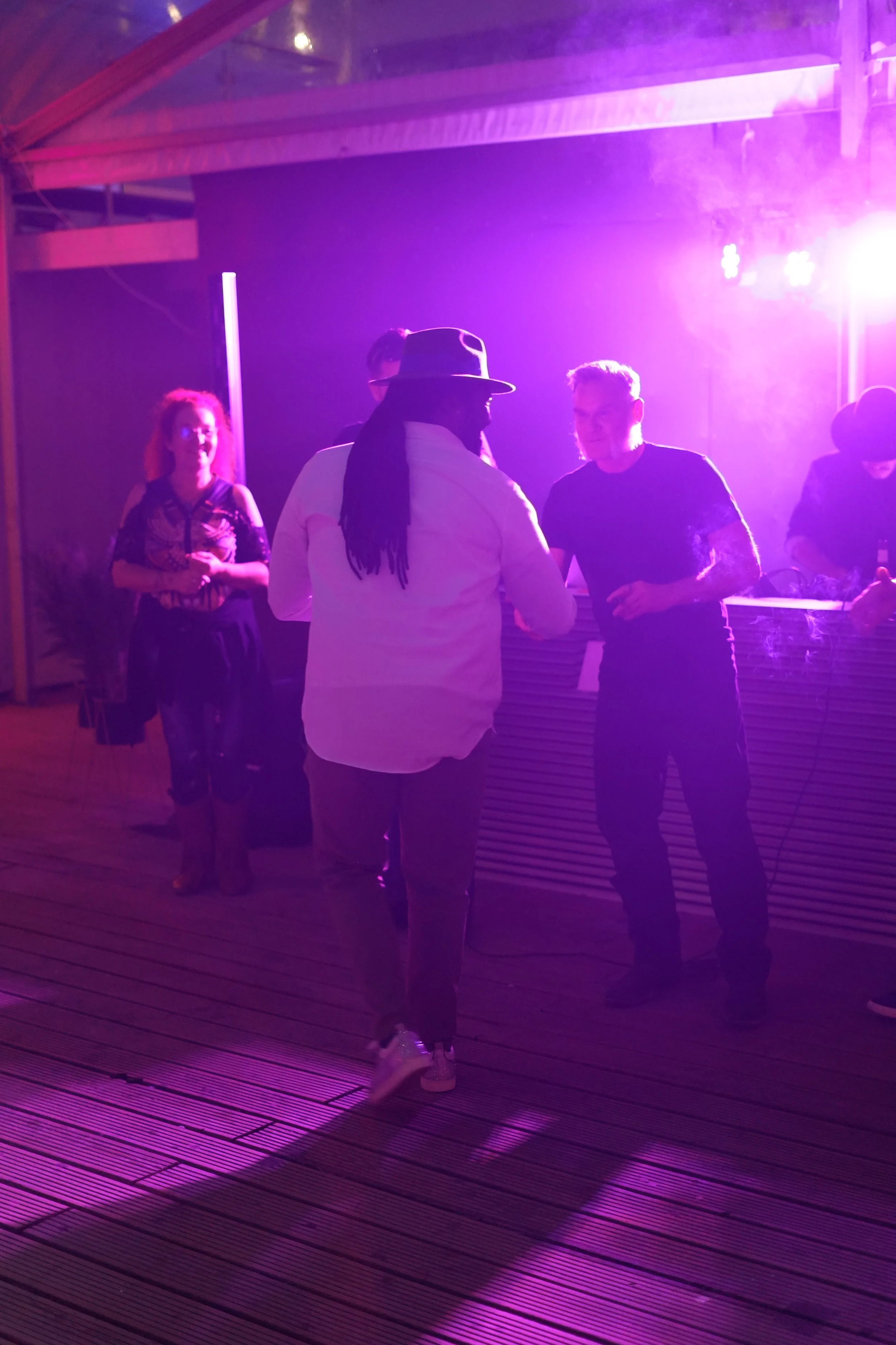 Group of people socializing at a nightclub with purple lighting, wooden flooring, and DJ setup.