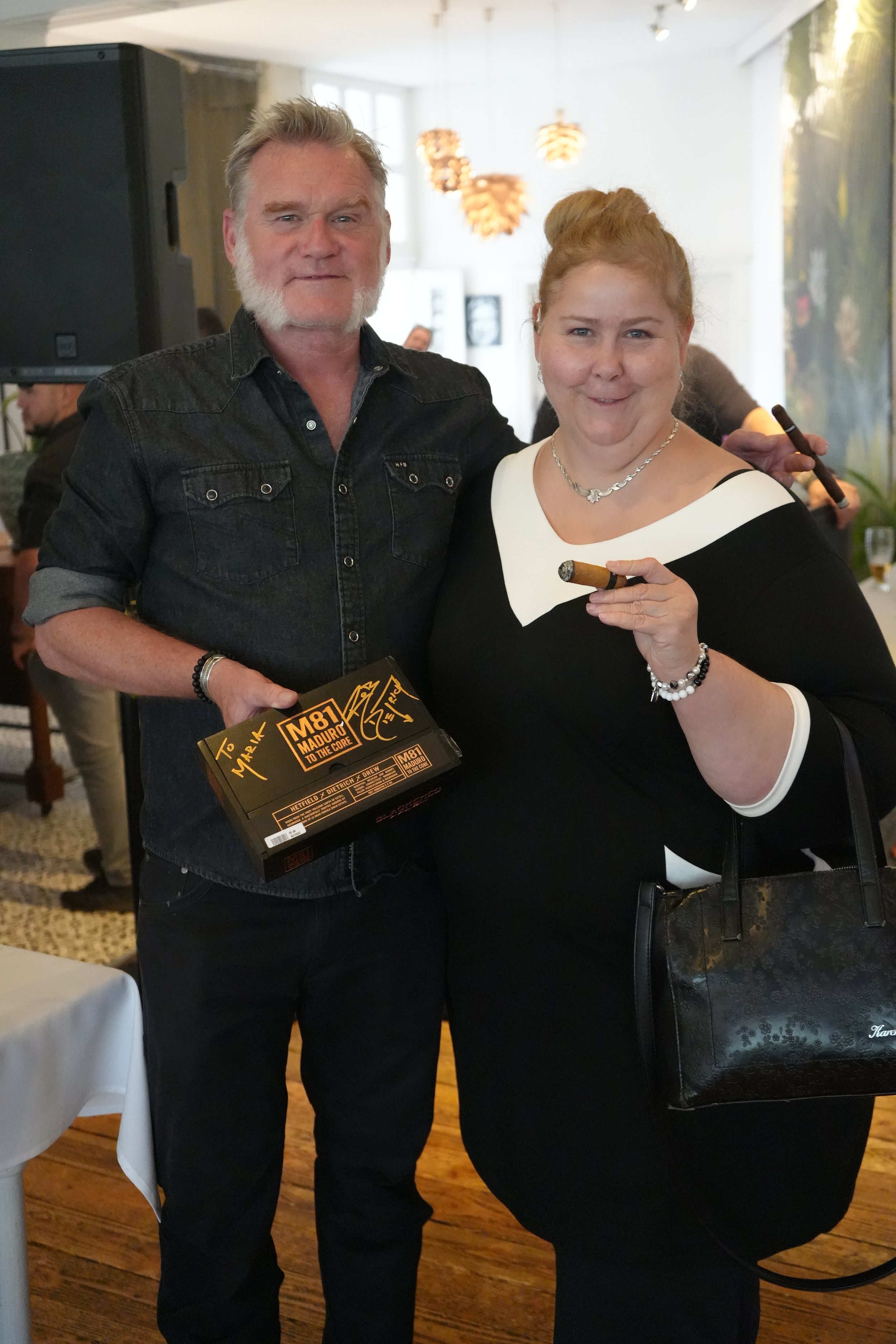 Two people standing together at a social event, one holding a box with a signed message. The man has gray hair and a beard, dressed in a black denim shirt, and the woman has red hair styled in a bun, wearing a black and white dress and holding a ciga