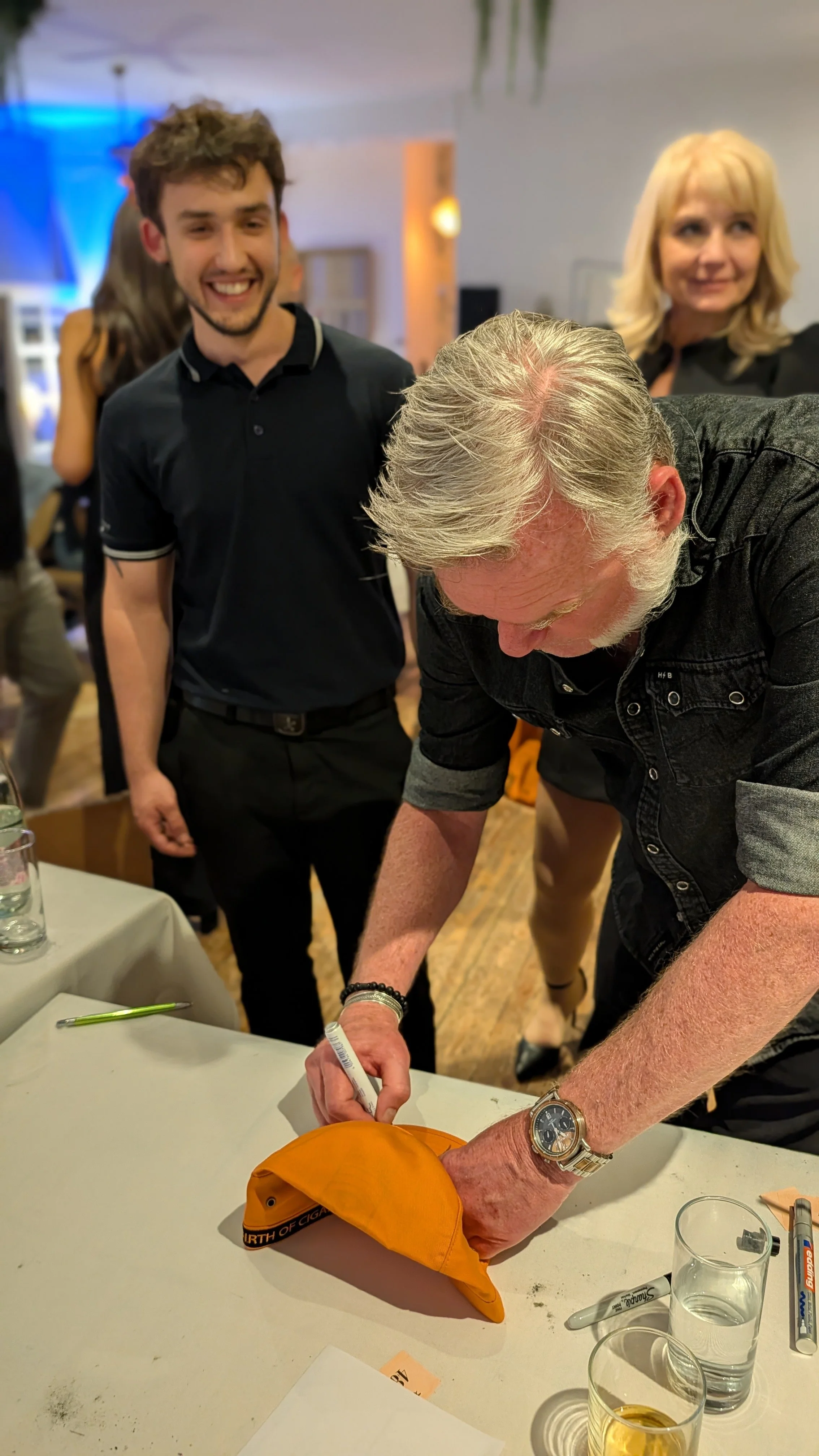 A group of people at a social gathering, with one man signing an orange cap on a table. Other individuals in the background are smiling and observing. The table has water glasses, markers, and a pen.