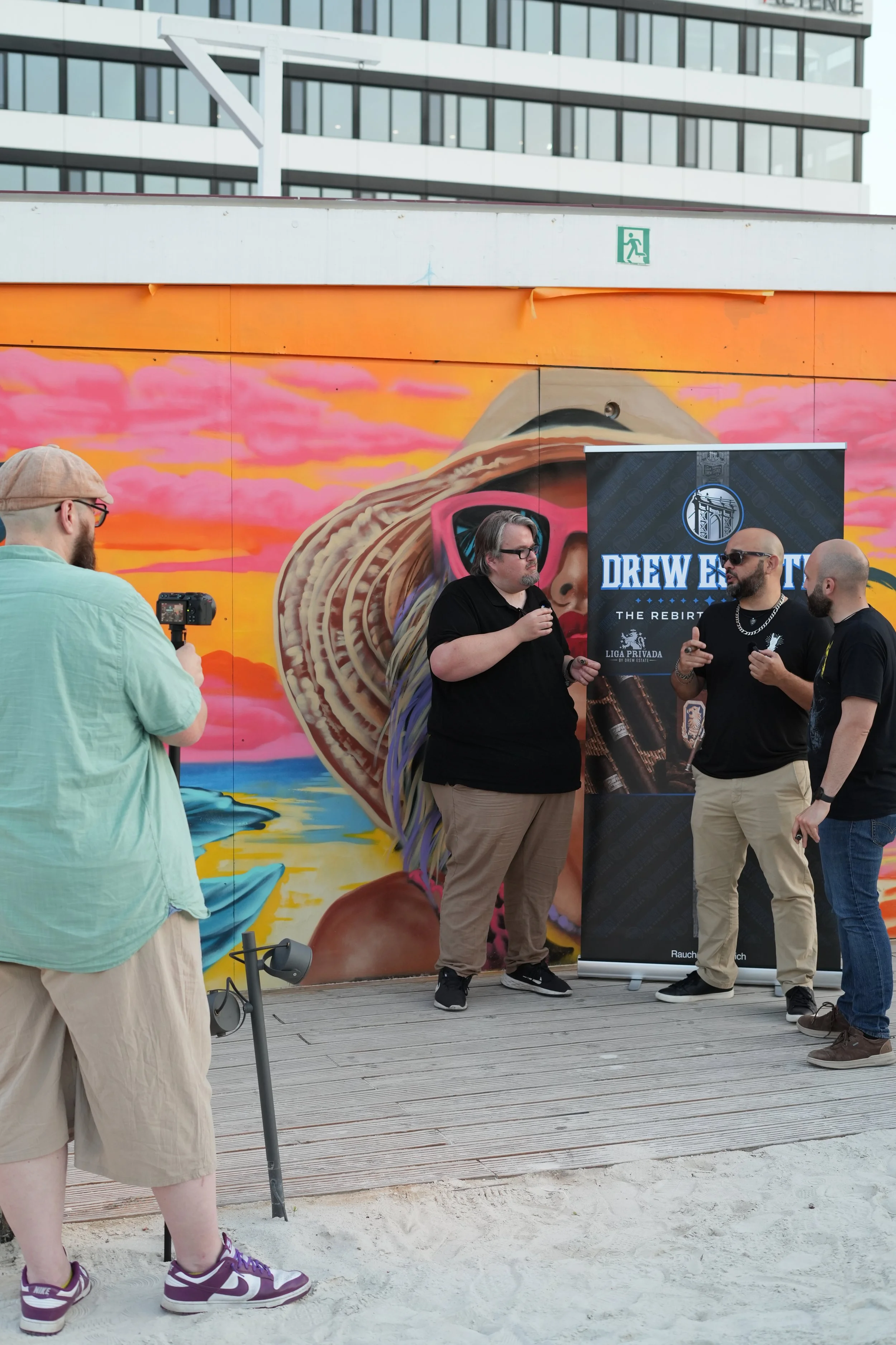 Four men are standing and talking in front of a colorful mural and a banner that reads "Drew Estate The Rebirth." One man is holding a microphone, while another is holding a camera. A fourth man has a shaved head, sunglasses, and a chain necklace.