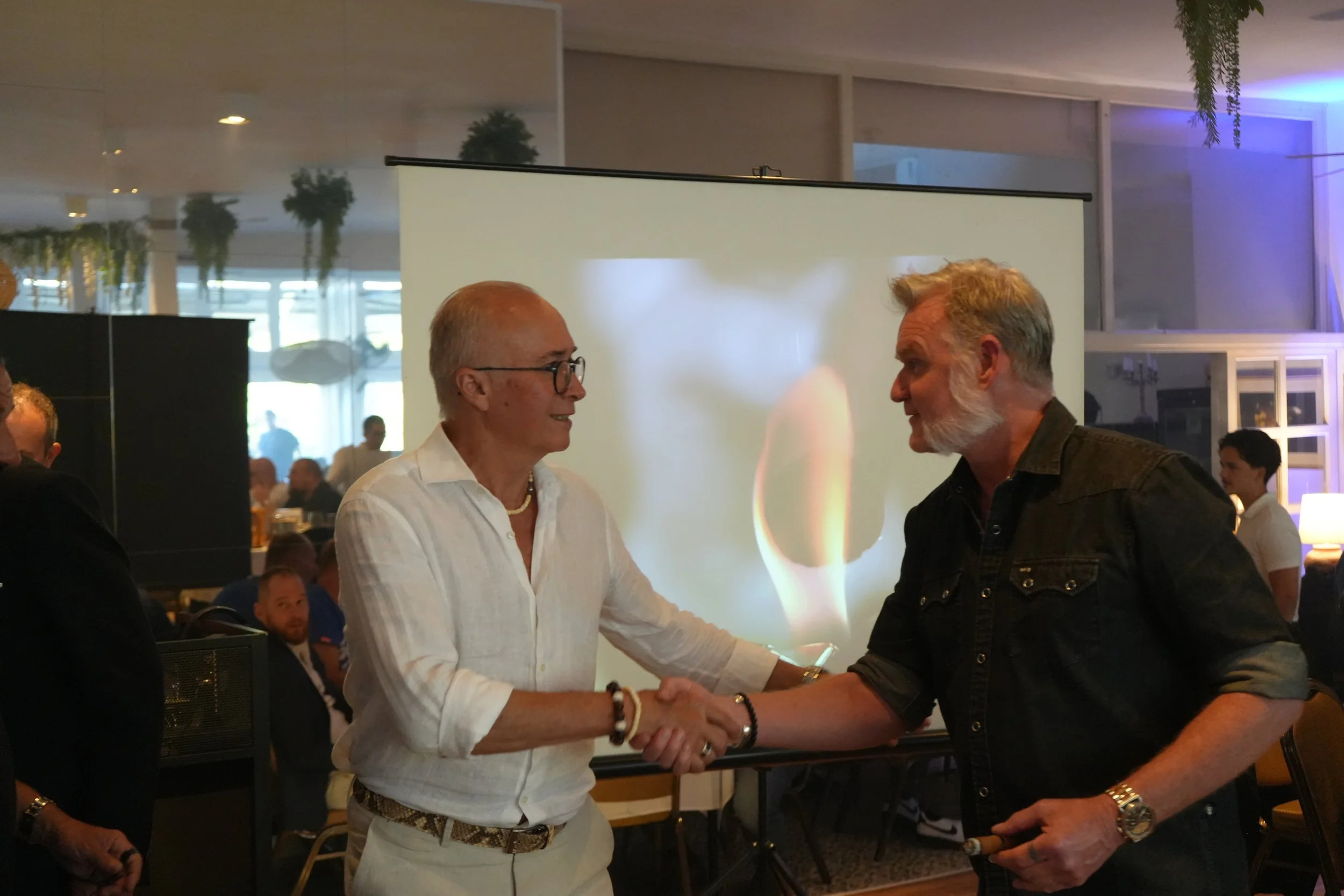 Two men shaking hands and smiling at each other in a social setting, with multiple people and a window in the background.