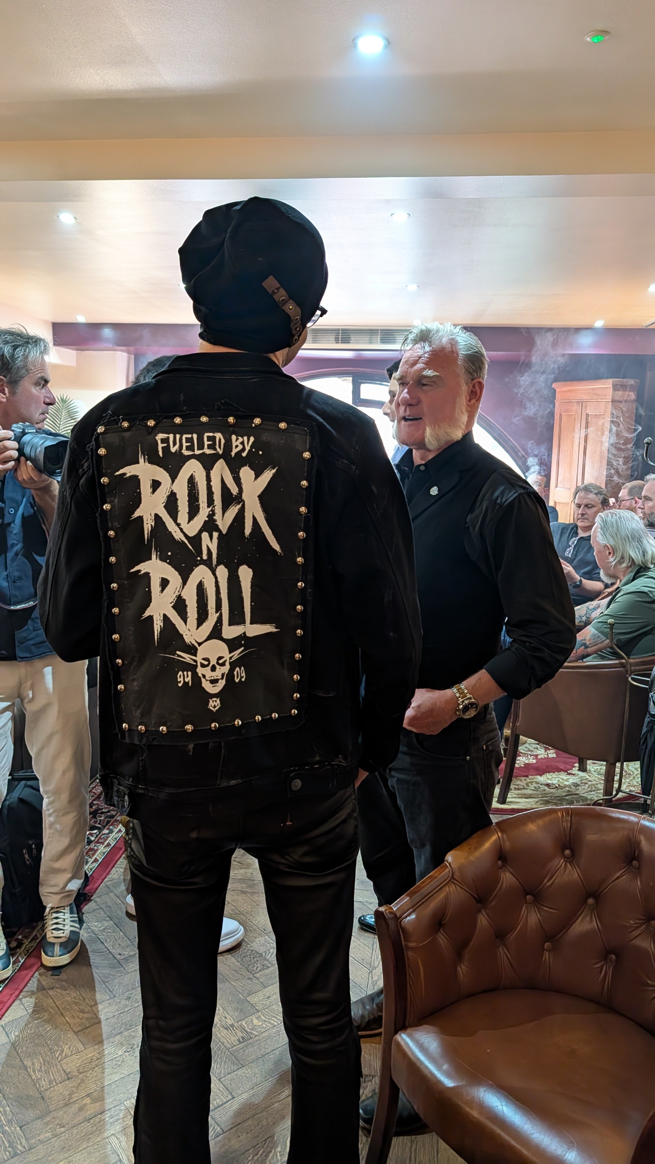 A man with sideburns and gray hair talking to another man wearing a black beanie and a black leather jacket with a painted logo that says 'Fueled by Rock n Roll' and a skull design.