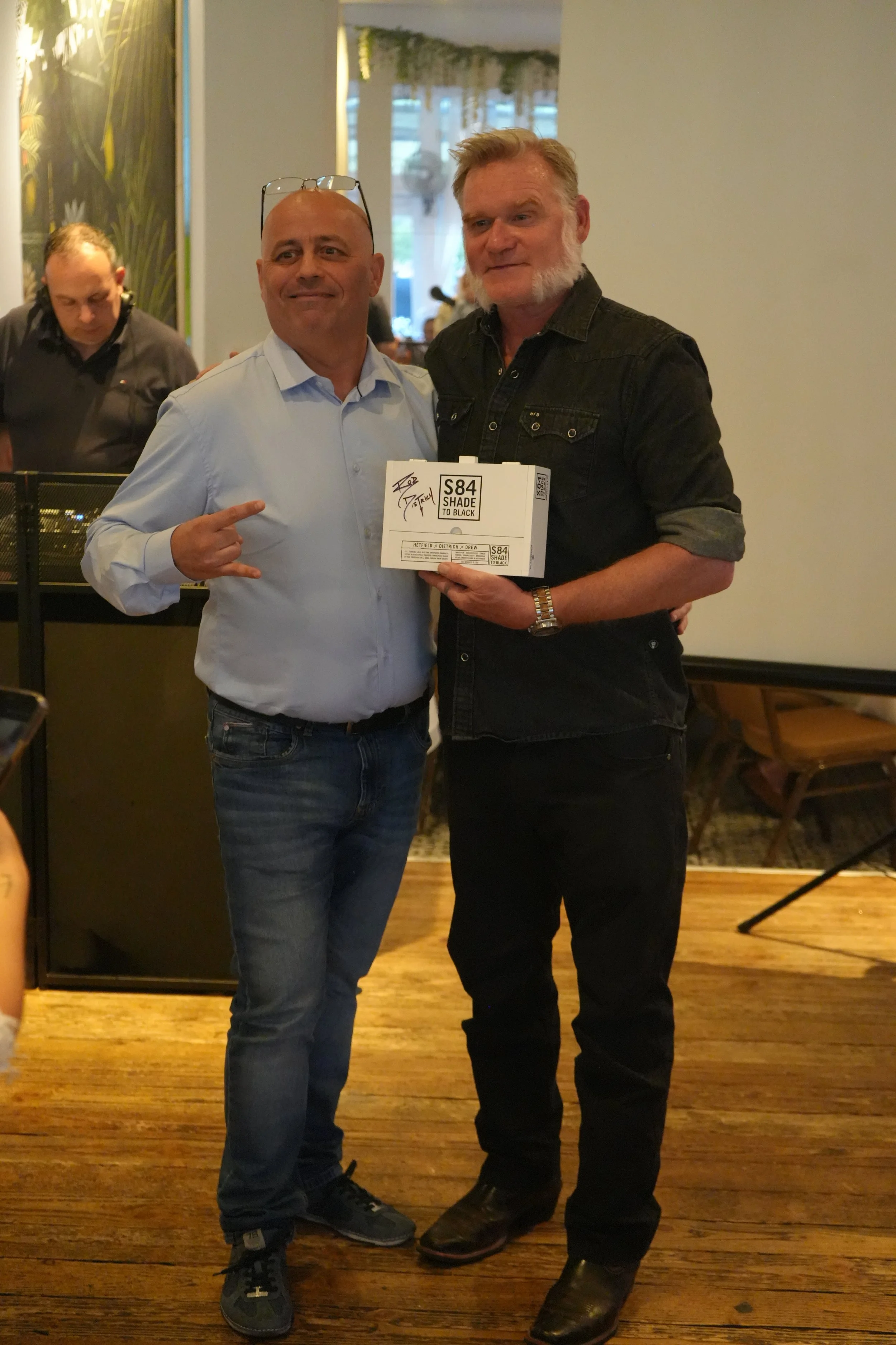 Two men standing indoors, one pointing to a box held by the other. The man on the left prefers casual clothing with a white shirt and jeans, while the man on the right wears a black denim shirt and boots. The man on the right is holding a box labeled