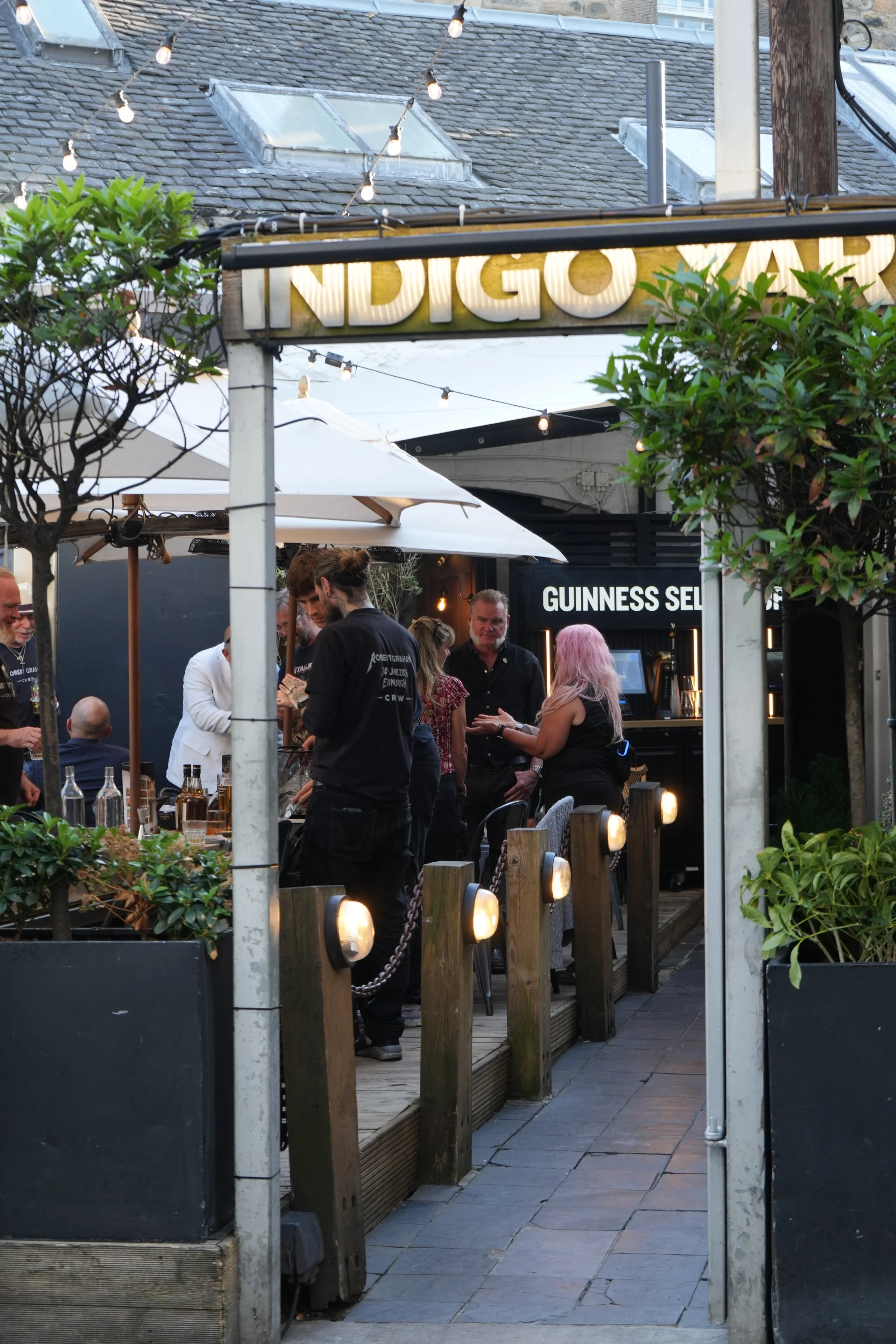 People gathering at an outdoor bar or restaurant, with a sign that partly reads 'INDIGO T...' and string lights hanging overhead.