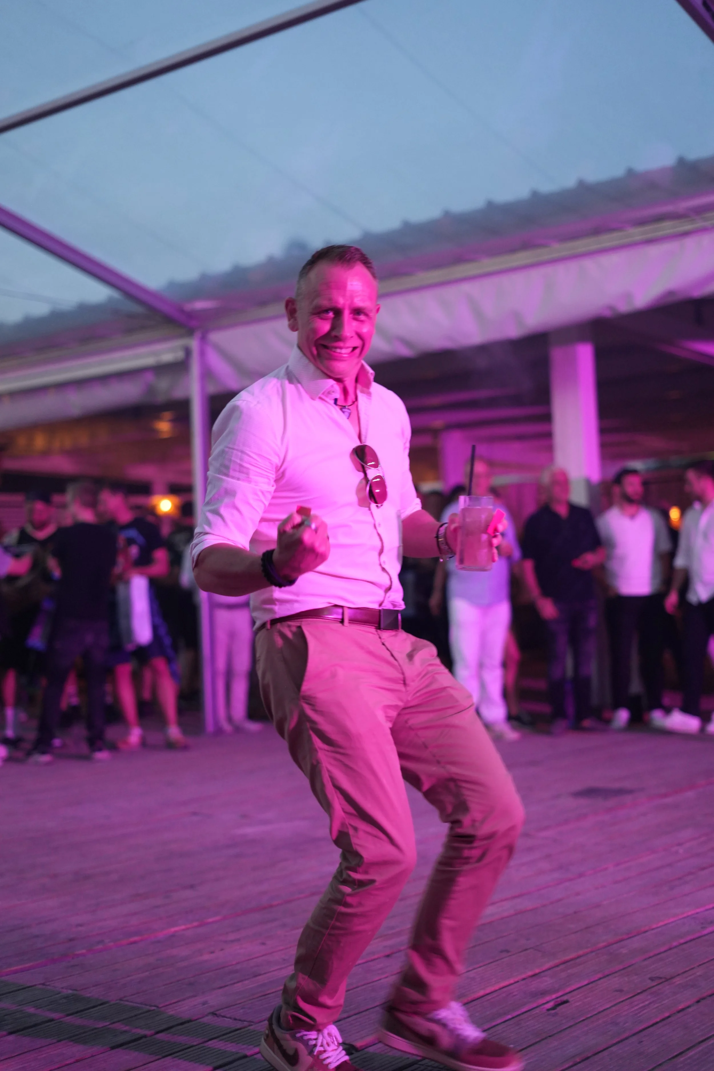 A man in light-colored pants and a white shirt dancing at an indoor event, holding a drink, with people standing and socializing in the background under purple and pink lighting.