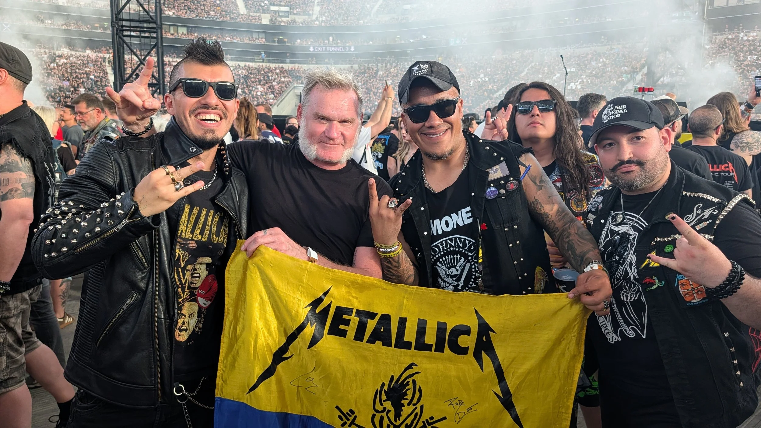 Group of people at a Metallica concert holding a yellow Metallica banner, some making rock hand signs, with a crowd and stage in the background.