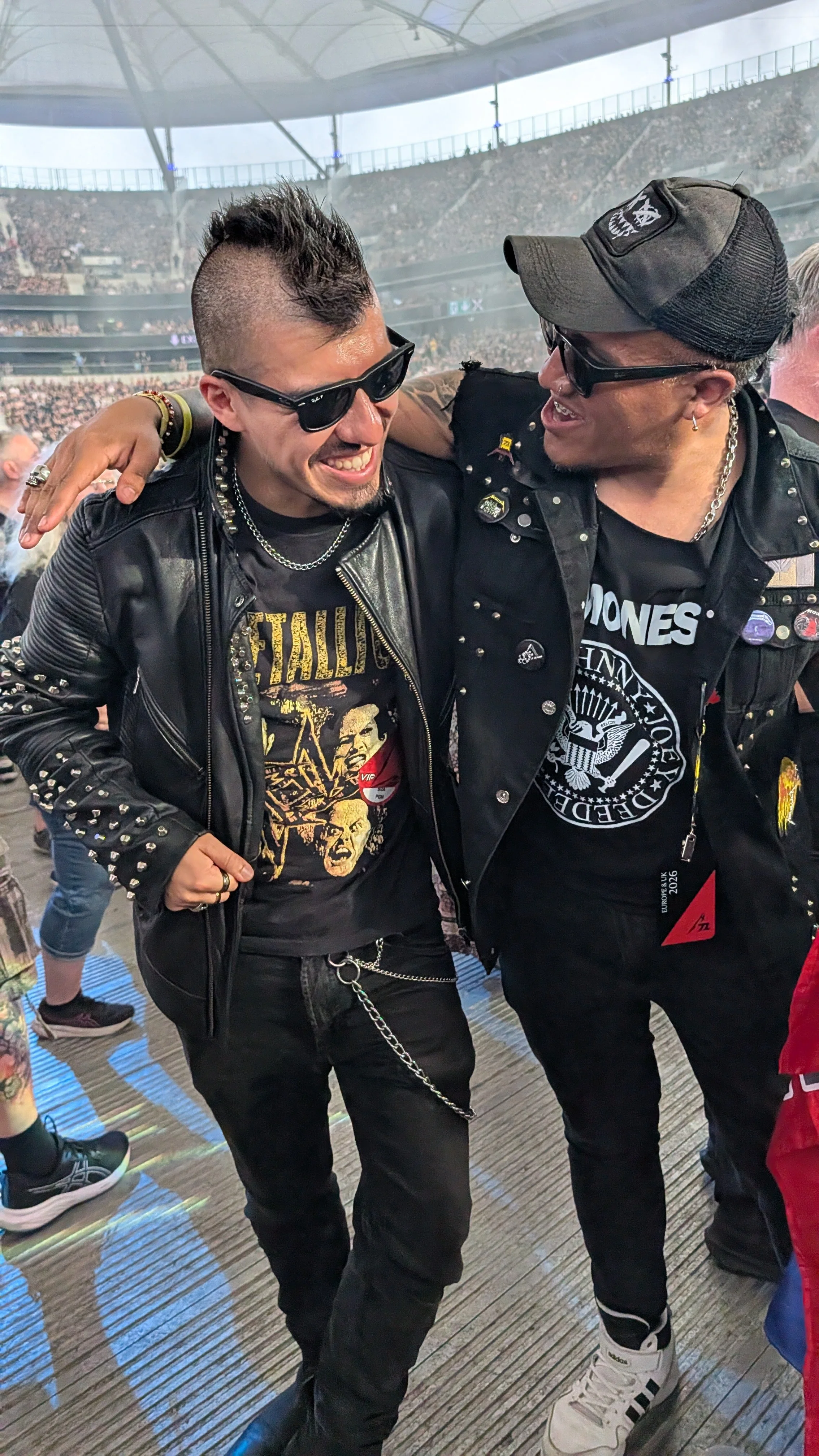 Two men in black leather jackets and T-shirts, one with a Metallica shirt and the other with a Ramones shirt, are smiling and embracing at a concert venue filled with a large crowd and a white canopy roof.