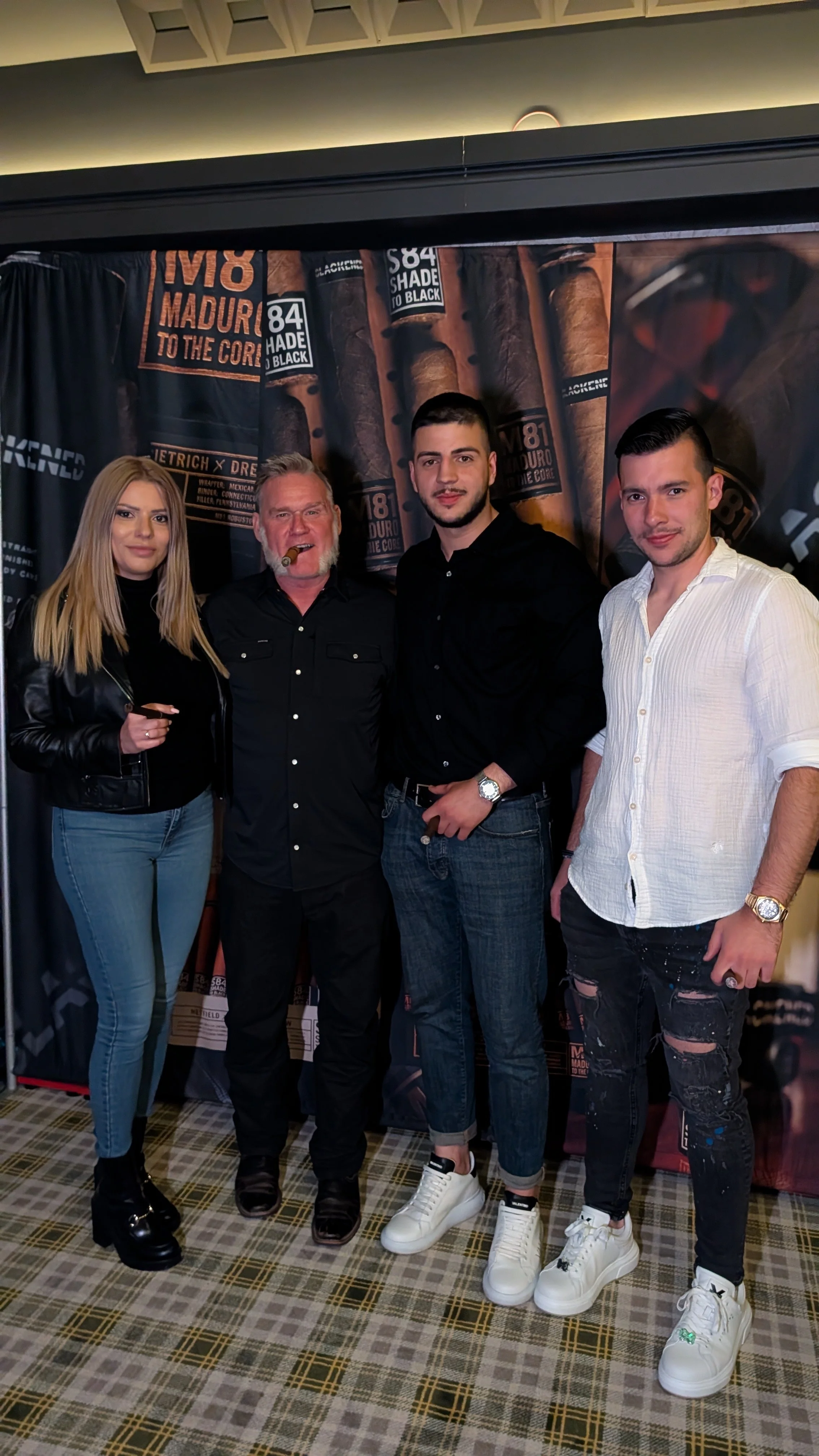 Four people stand together in front of a cigar-themed backdrop, holding drinks and posing for the photo. They are in a room with plaid carpet and subdued lighting.