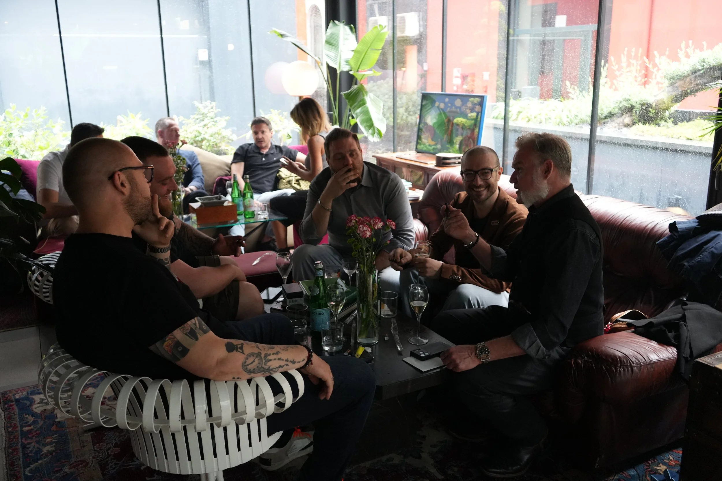 Group of people sitting at a table in a restaurant with large windows and indoor plants, some engaging in conversation and others using their phones.