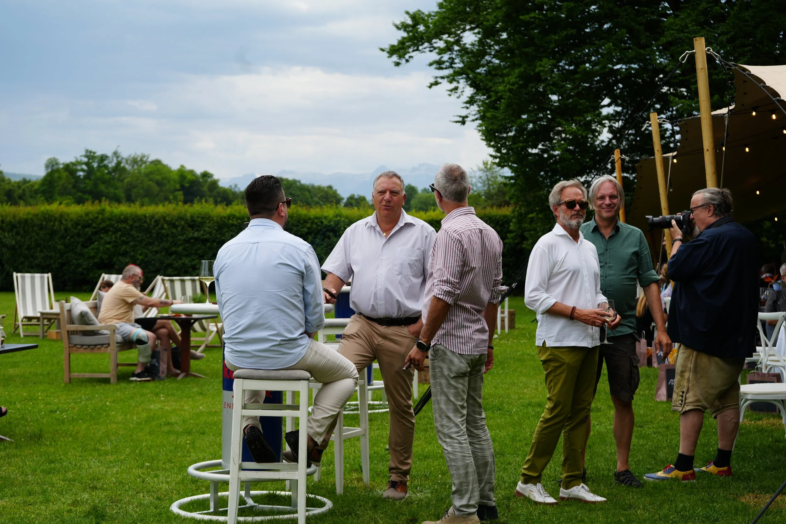 A group of men socializing outdoors on a lawn during daytime, with some sitting on chairs and others standing, under a cloudy sky with trees in the background, and a tent with string lights on the right.
