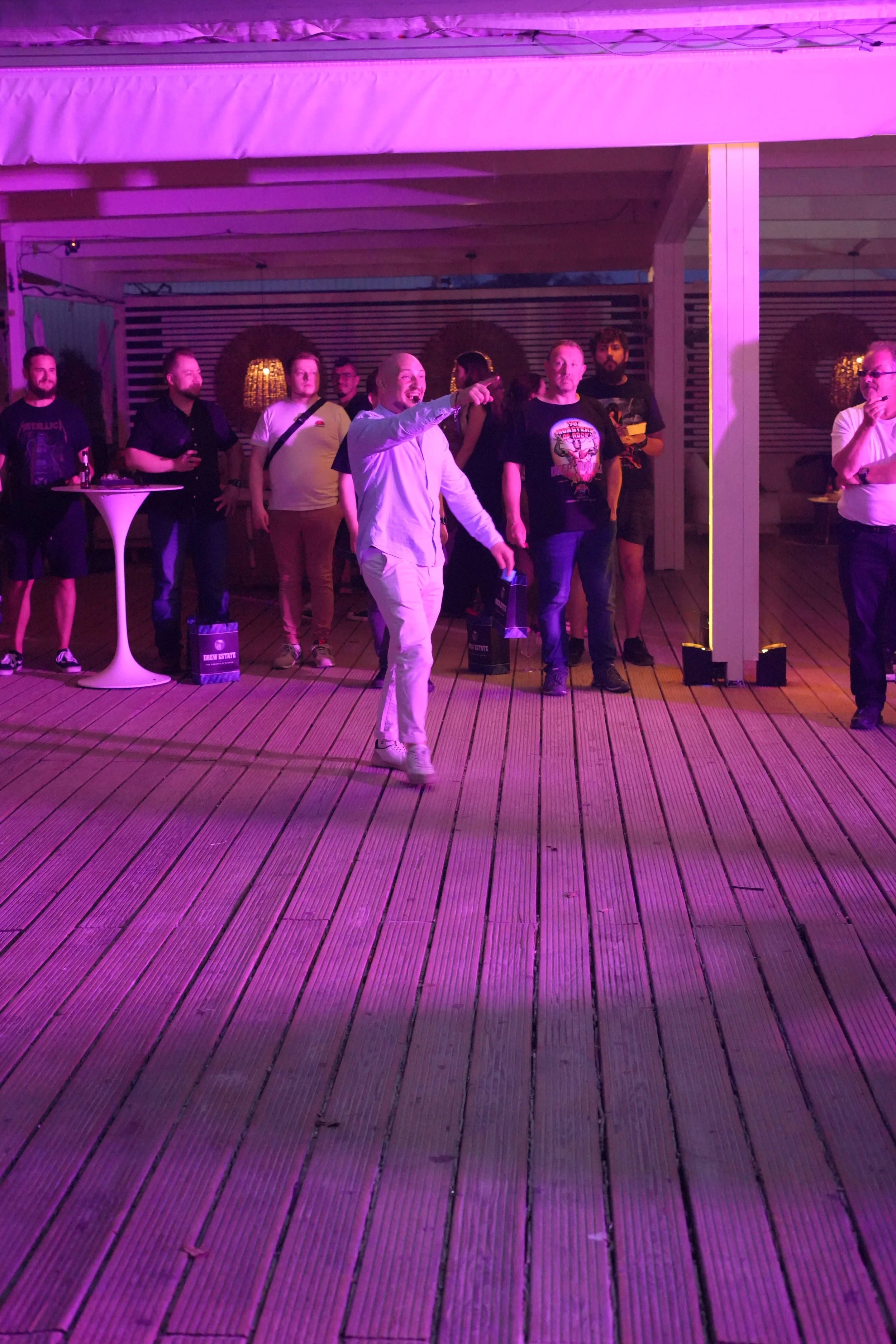 People gathered on a wooden deck under purple lighting, with some individuals holding drinks and engaging in conversation at a social event.