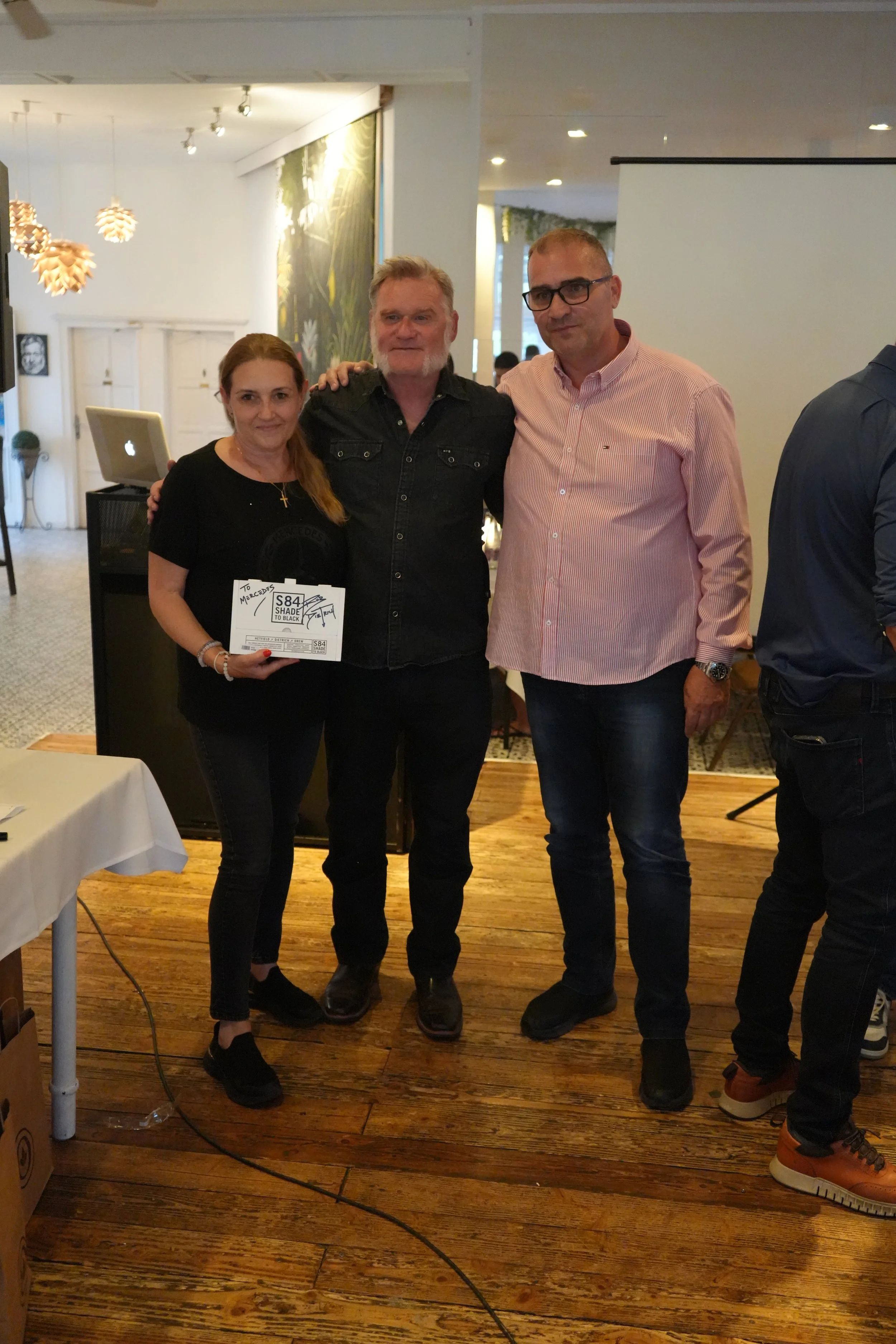 Three people standing together indoors, smiling at the camera. The woman on the left is holding a sign that says 'To Mercedes,' and is wearing black clothing and shoes. The man in the middle has grey hair and a beard, wearing a black denim shirt. The